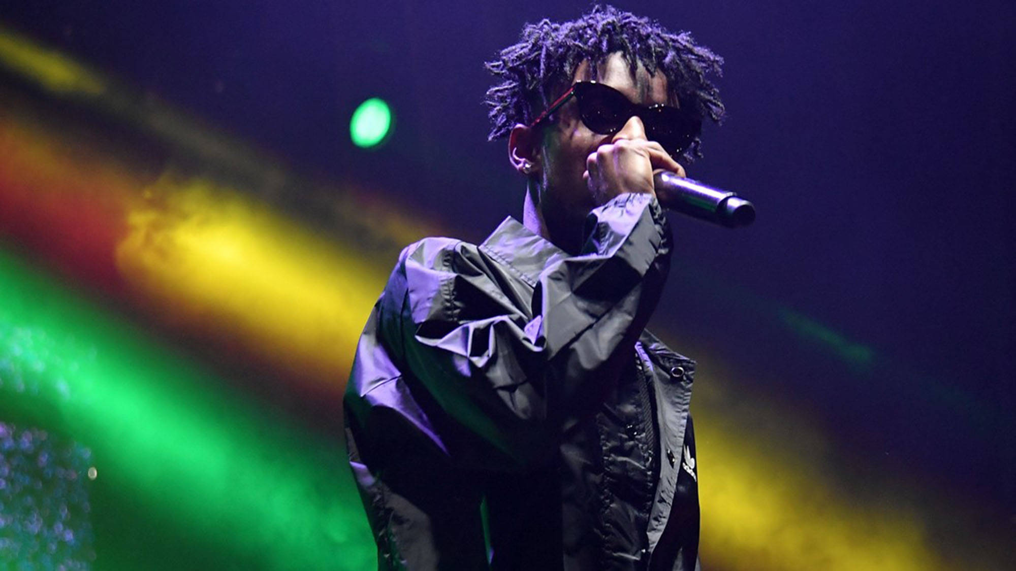 21 Savage Adidas Performance 壁紙 — 一位梳著辮子頭、戴著太陽眼鏡的饒舌歌手，在充滿活力的綠色、黃色和紫色舞台燈光下對著麥克風表演。