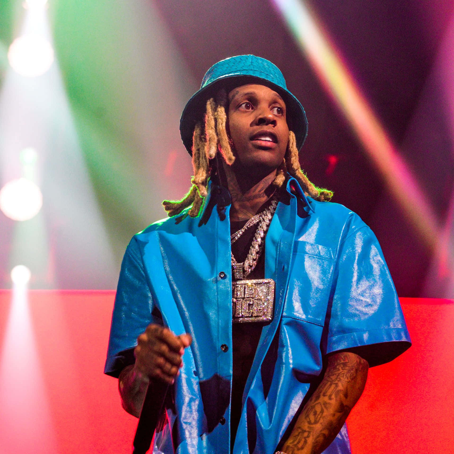 lil durk 桌布 壁紙 — 一位梳著辮子頭、戴藍色帽子的說唱歌手在彩色聚光燈下表演，身穿閃亮藍色襯衫和厚重珠寶。