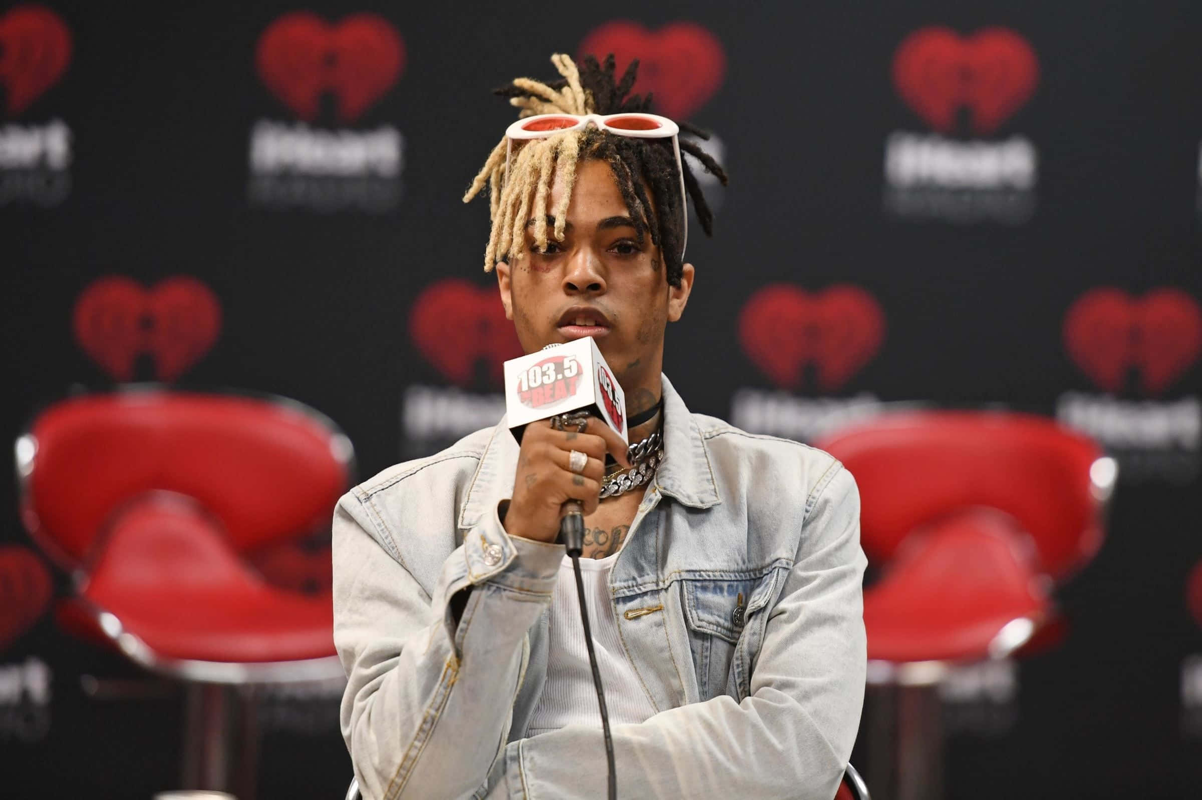XXXTENTACION 桌布 壁紙 — 一位梳著辮子頭、頭上戴著紅色太陽眼鏡的饒舌歌手在iHeartRadio活動中對著麥克風說話，身後有紅色椅子和心形標誌。