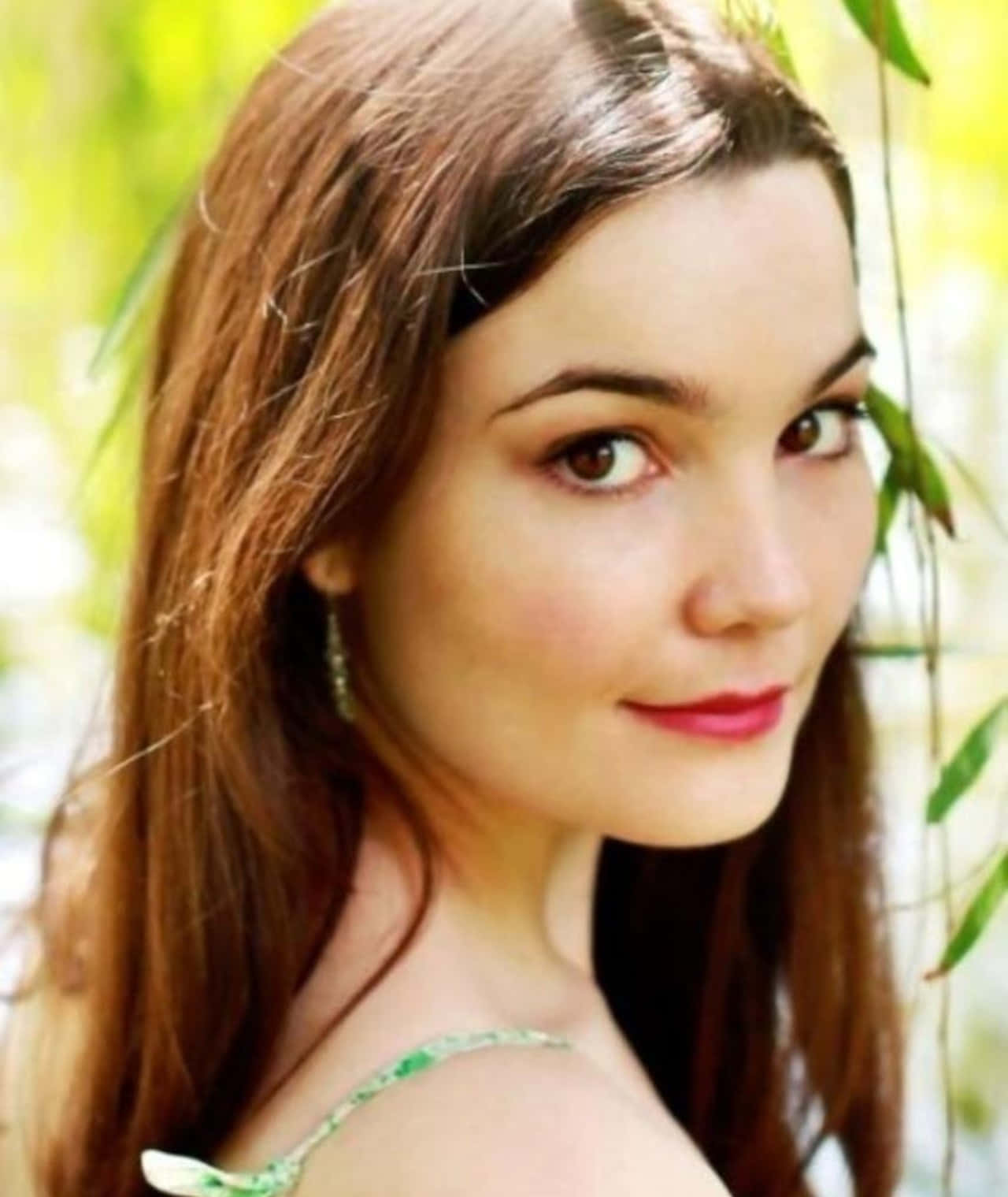 Bridie Latona 桌布 壁紙 — 一位棕色頭髮、塗著紅色唇膏的年輕女性，在明亮的綠色樹葉背景前溫柔地微笑。