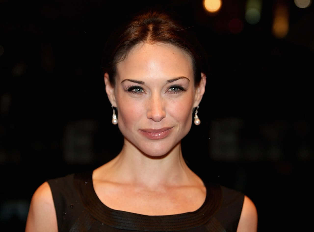 Claire Forlani 桌布 壁紙 — 一位棕色頭髮、戴著珍珠耳環的女性，身穿黑色連衣裙，在溫暖散景燈光下的模糊深色背景中溫柔微笑。