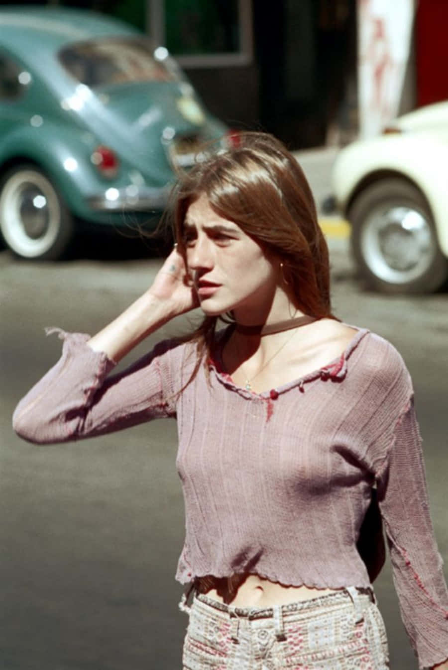70年代gif 桌布 壁紙 — 一位棕色頭髮的年輕女子站在陽光普照的街道上，手靠近耳朵，身穿粉紅色短款毛衣和圖案褲子，背景中有模糊的復古汽車。