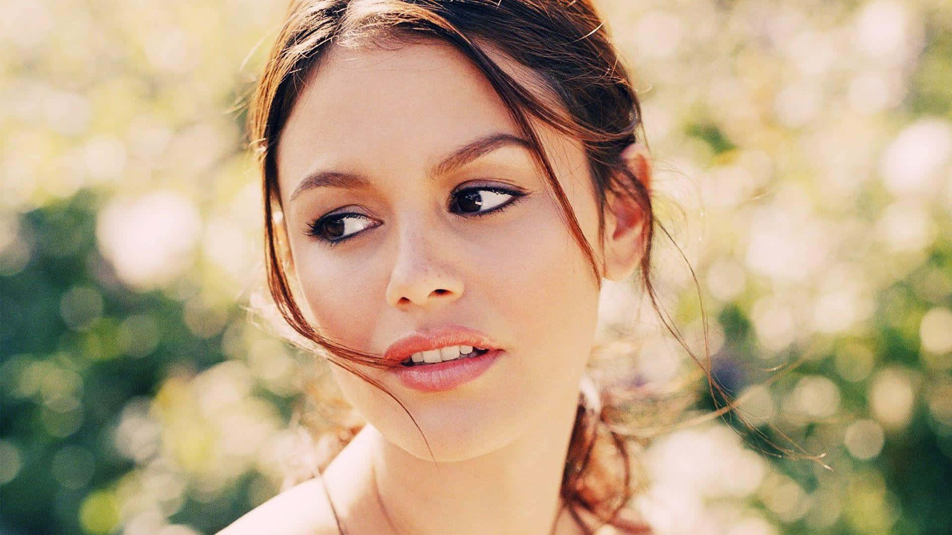 Rachel Bilson 桌布 壁紙 — 一位棕色頭髮的年輕女性在陽光下，對著模糊的綠色和白色花卉背景，若有所思地望向一旁。
