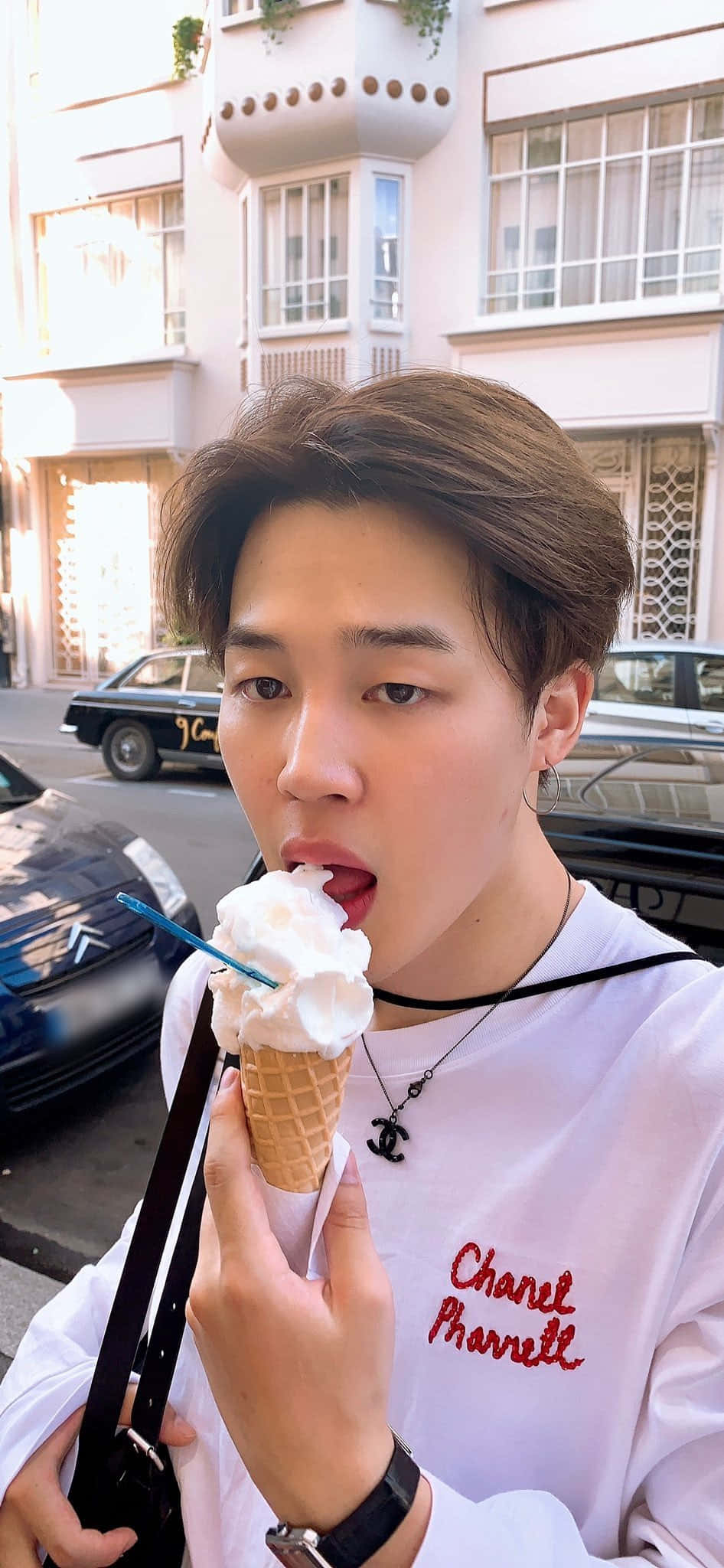 Jimin Aesthetic 桌布 壁紙 — 一位棕色頭髮的年輕男子在陽光明媚的城市街道上享用雲呢拿雪糕筒，身後有復古汽車和粉彩建築。