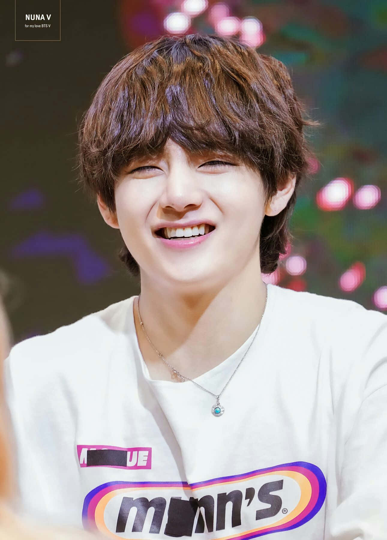 Taehyung Cute 桌布 壁紙 — 一位棕色頭髮的年輕男子穿著白色圖案T恤，在色彩繽紛的模糊背景前露出燦爛笑容。