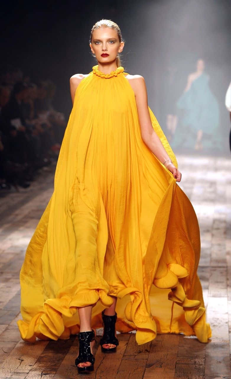 Yellow Fashion 桌布 壁紙 — 一位模特兒在戲劇性的舞台燈光下，自信地穿著一件寬大飄逸的黃色長裙走過天橋。