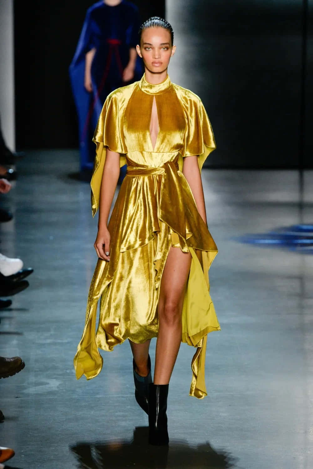Yellow Fashion 桌布 壁紙 — 一位模特兒在明亮的舞台燈光下，自信地穿著閃閃發光的金色緞面連衣裙，帶有高開衩和斗篷袖，走在天橋上。