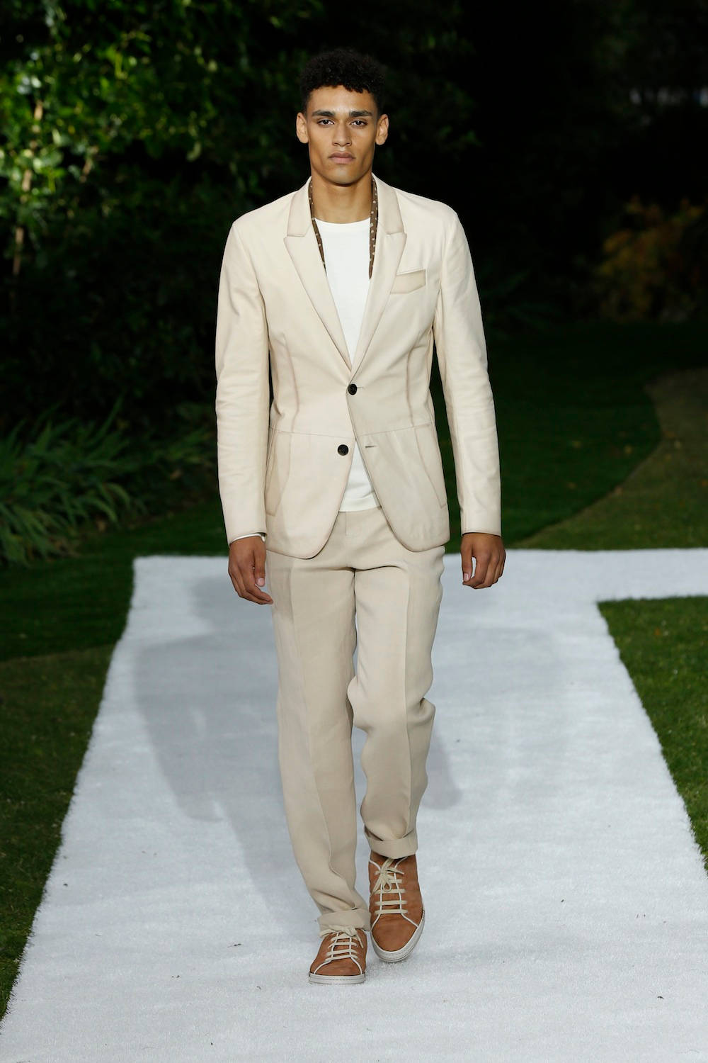 Berluti SS15 壁紙 — 一位模特兒自信地走在白色天橋上，周圍是茂密的綠植，在明亮的日光下。