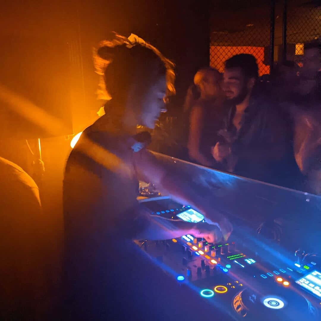 DJ 桌布 壁紙 — 一位正在移動的DJ在溫暖橙色和冷藍色的俱樂部燈光下操控發光的控制器，背後是模糊的人群。