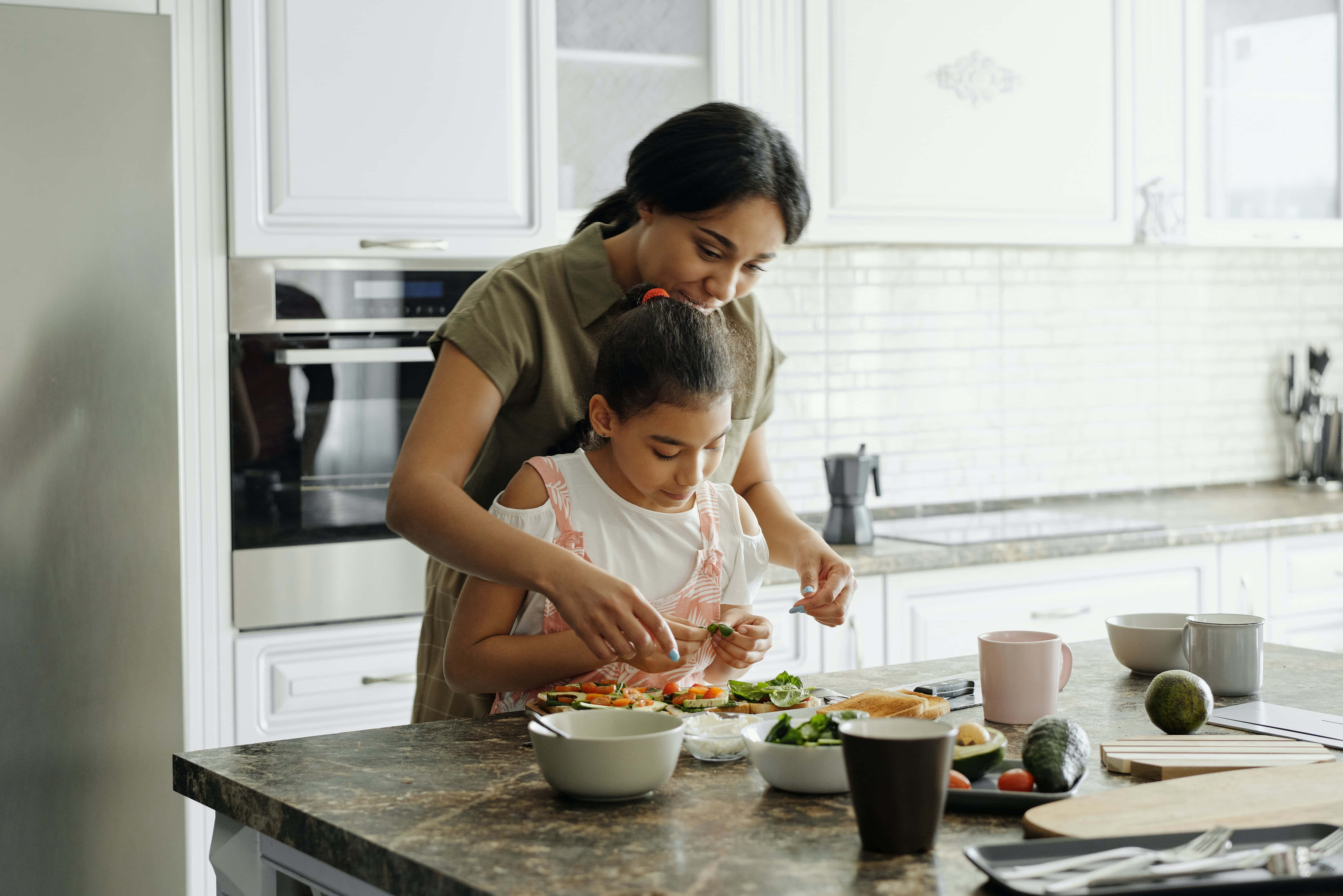 Mom 桌布 壁紙 — 一位母親和女兒在一個明亮現代、配有白色櫥櫃和花崗岩檯面的廚房裡一起準備色彩繽紛的沙律。