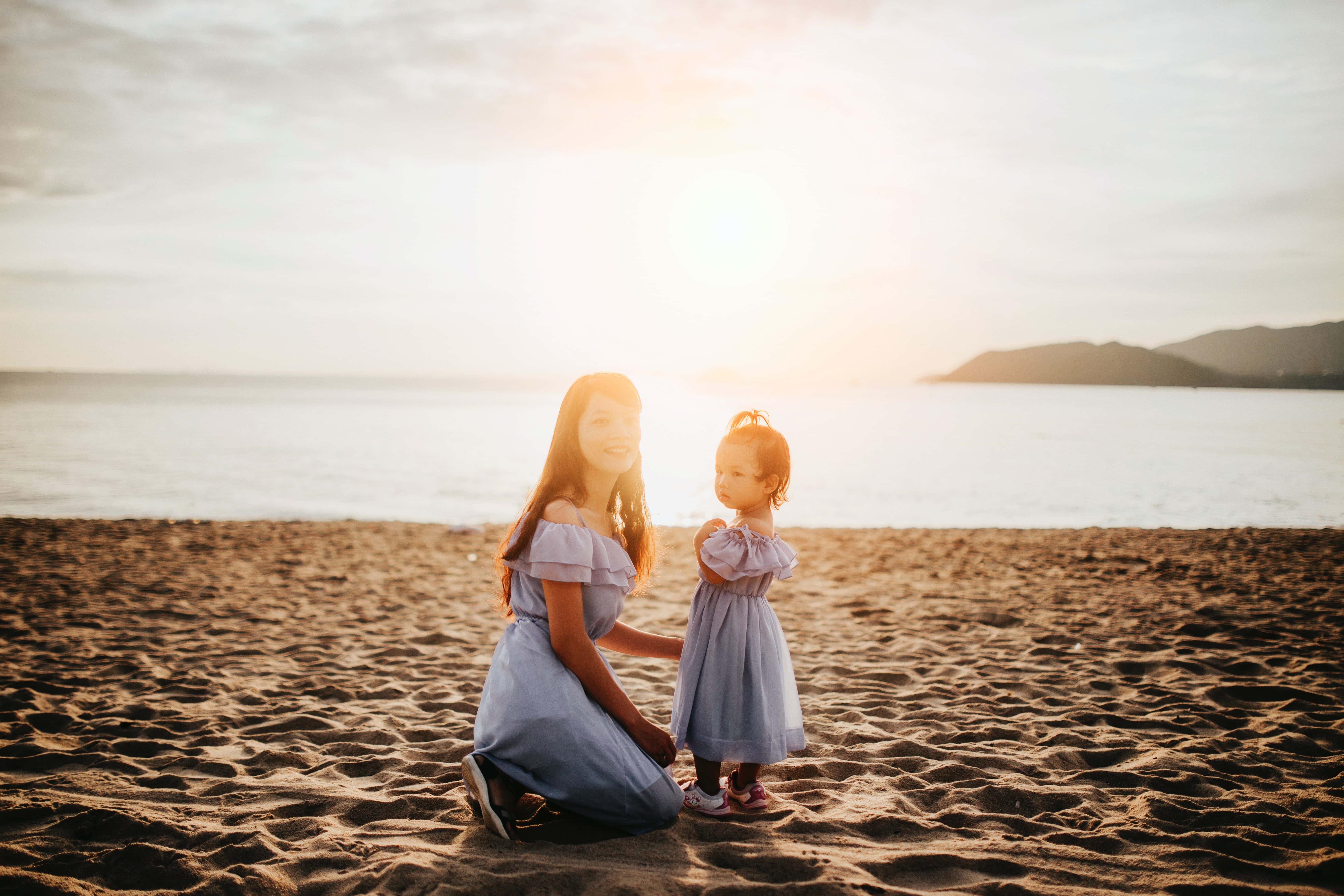 Mom 桌布 壁紙 — 一位母親和女兒穿著相配的藍色連衣裙，在日落時分的陽光沙灘上分享溫馨時刻。