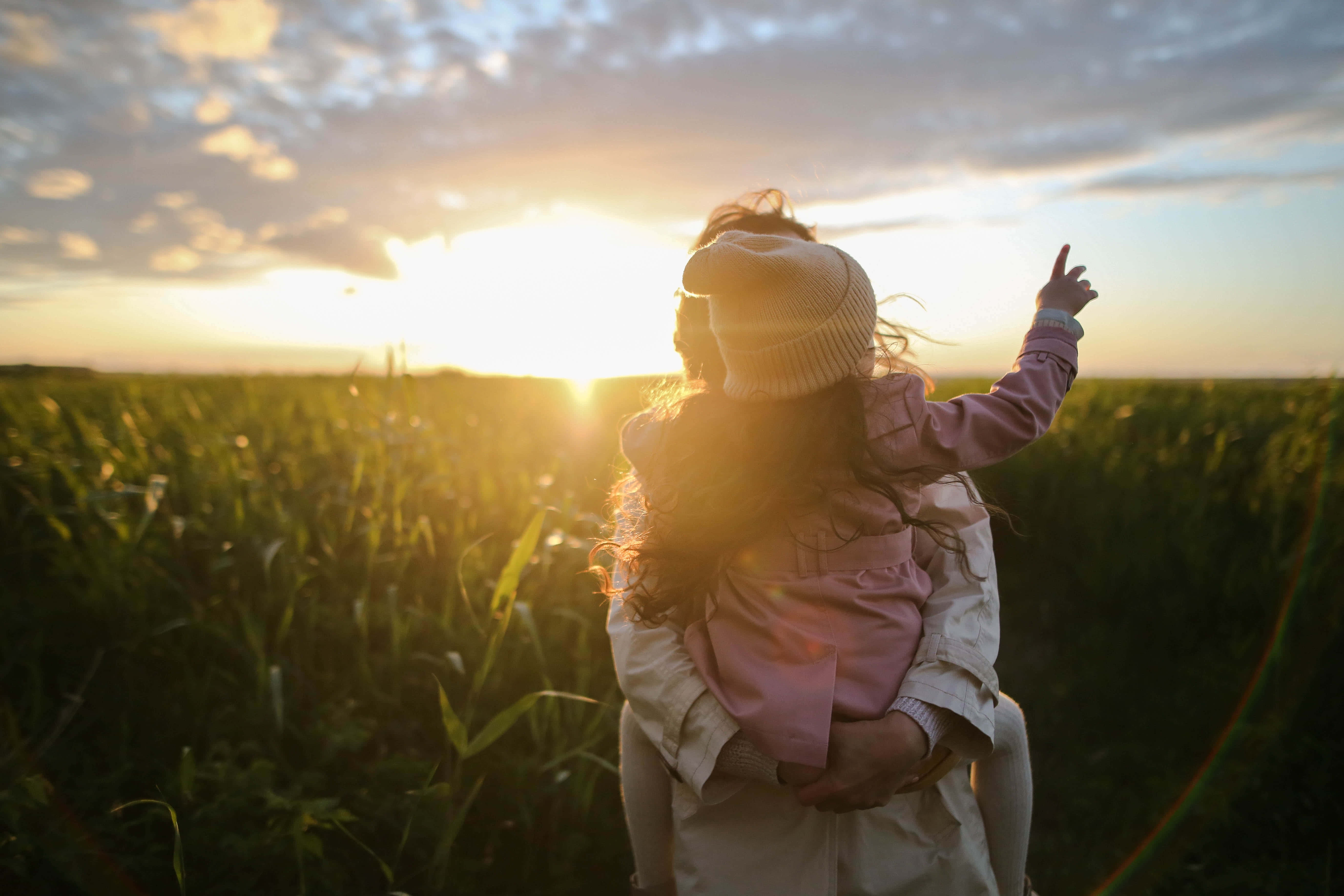 Mom 桌布 壁紙 — 一位母親在日落時分抱著孩子穿過陽光明媚的田野，孩子指向發光的地平線。