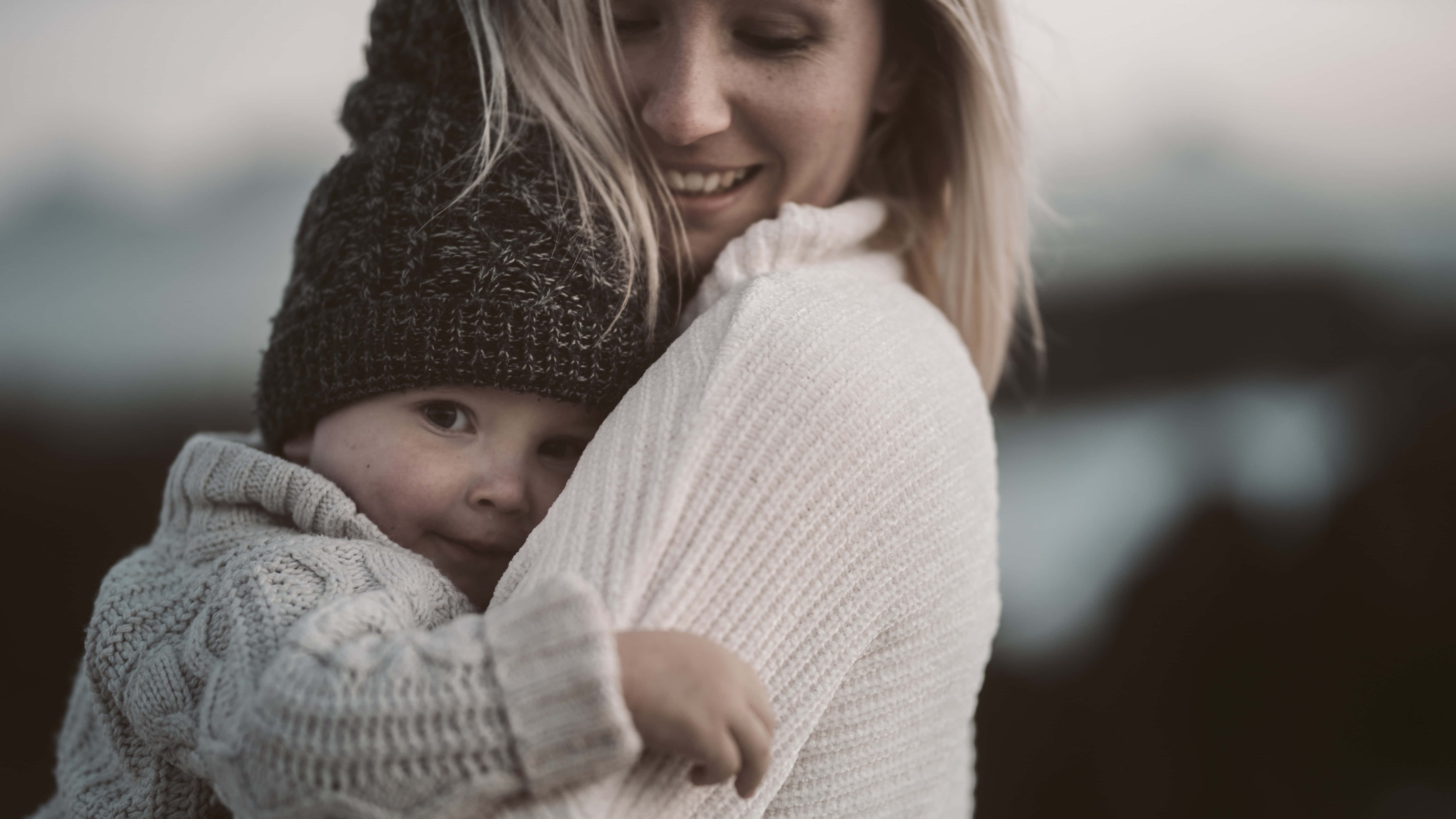 Mom 桌布 壁紙 — 一位母親溫暖地擁抱她的孩子，兩人都裹著舒適的針織毛衣，背景是柔和模糊的戶外景色。