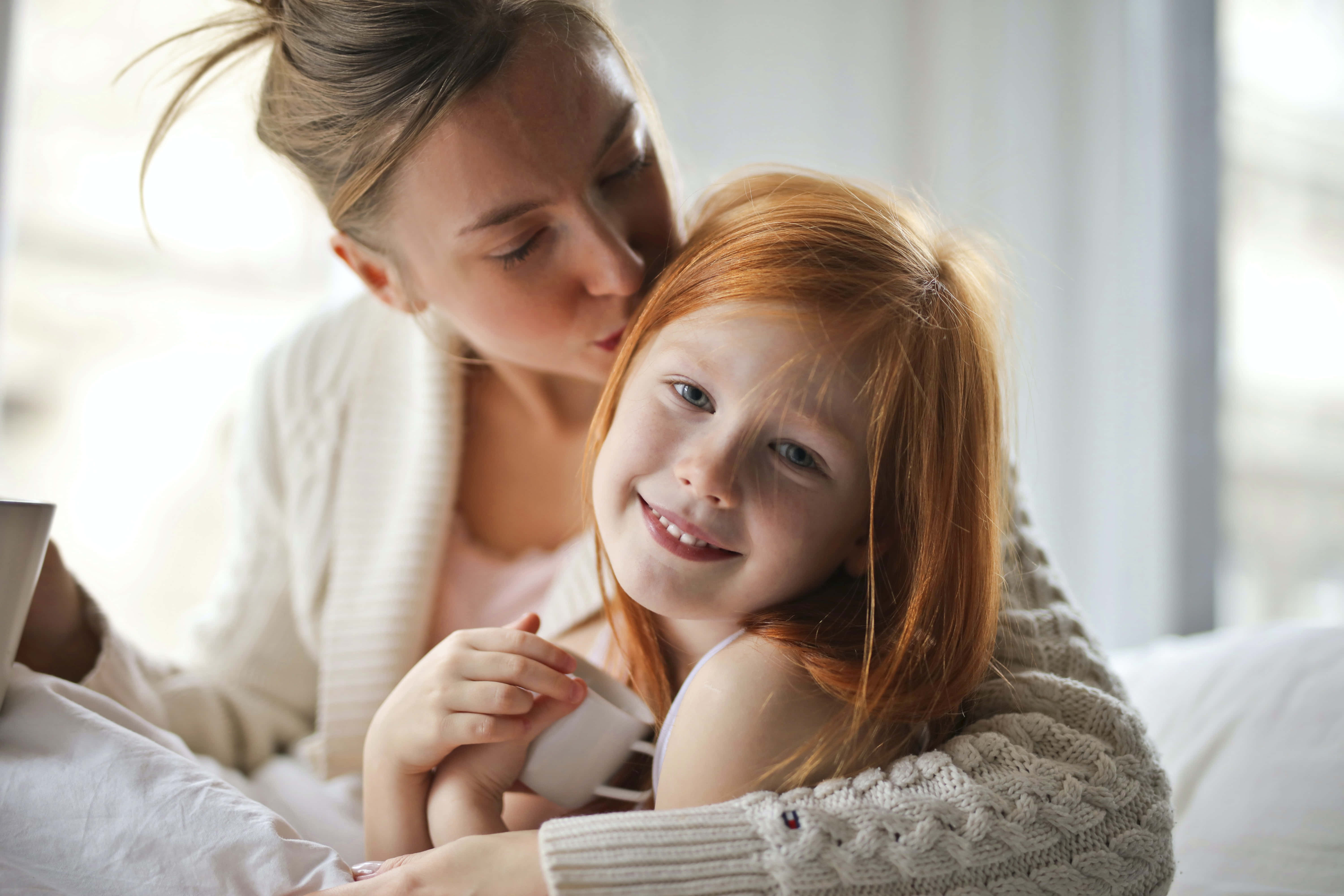 Mom 桌布 壁紙 — 一位母親溫柔地親吻她微笑的紅髮女兒的額頭，兩人舒適地坐在床上。
