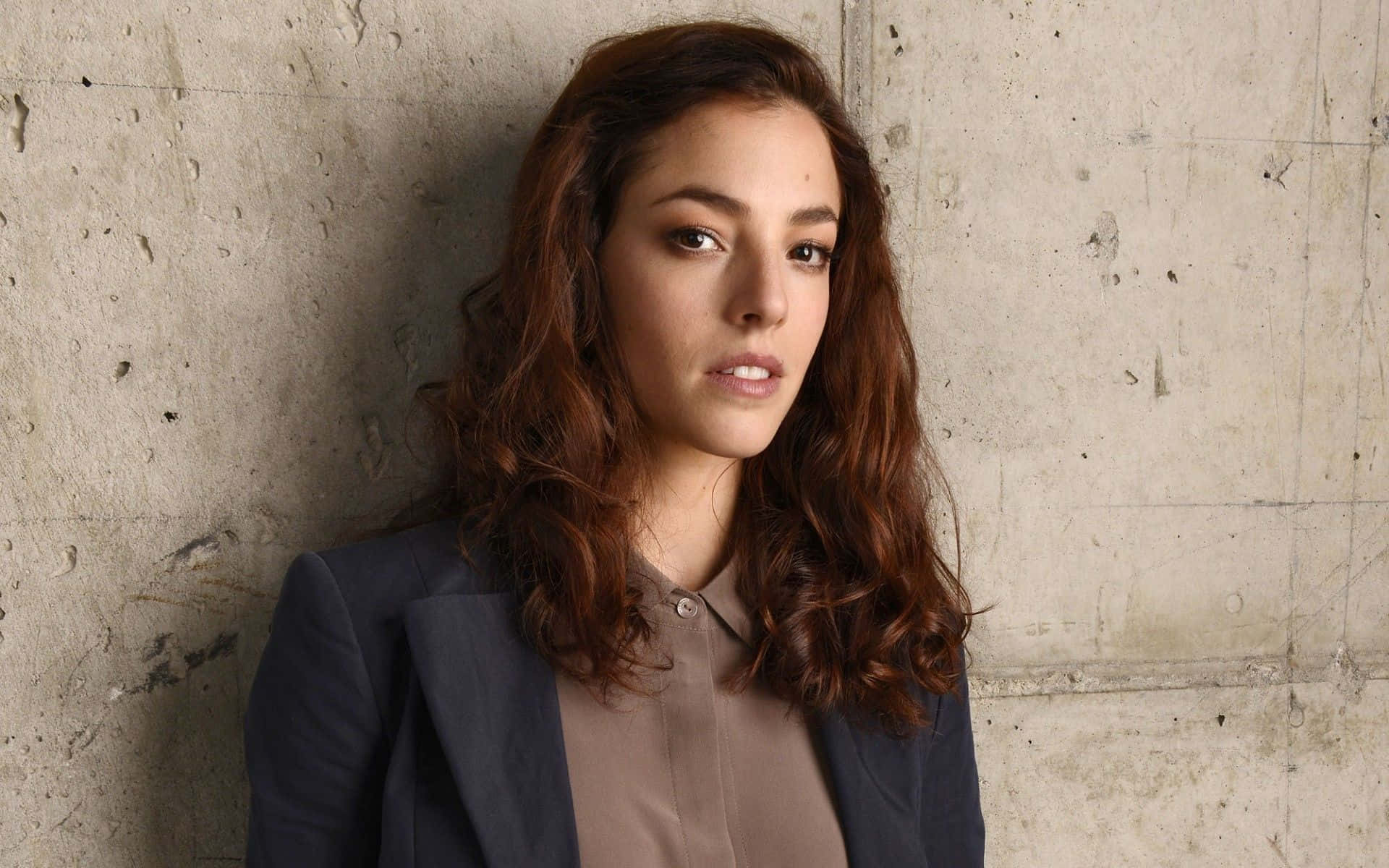 Olivia Thirlby 桌布 壁紙 — 一位波浪棕色頭髮的女性，身穿深色西裝外套和米色襯衫，靠在有紋理的混凝土牆上。