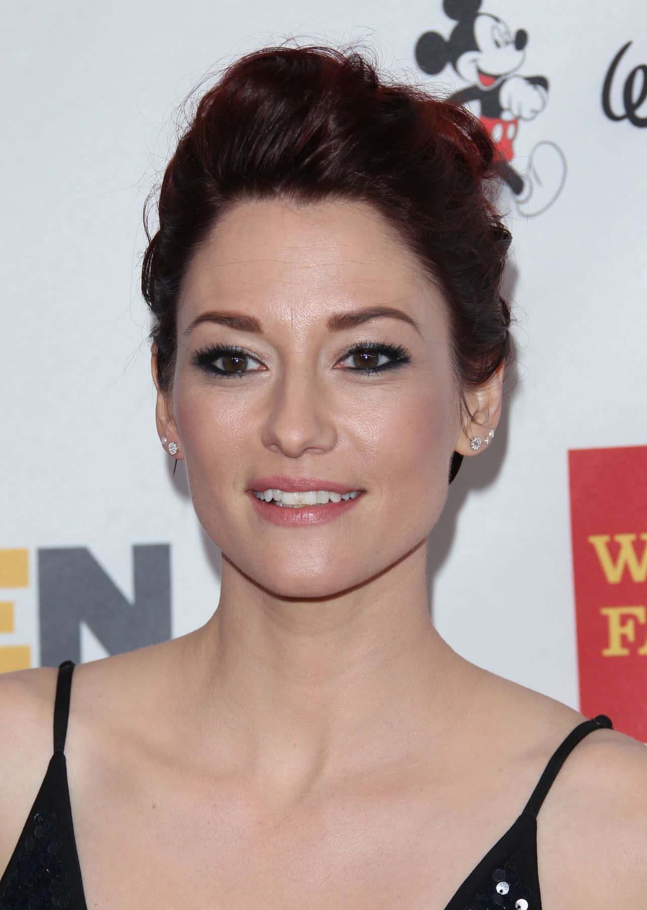 Chyler Leigh 桌布 壁紙 — 一位深紅色頭髮盤起的女性在迪士尼活動中穿著黑色連衣裙和鑽石耳環對著鏡頭微笑。