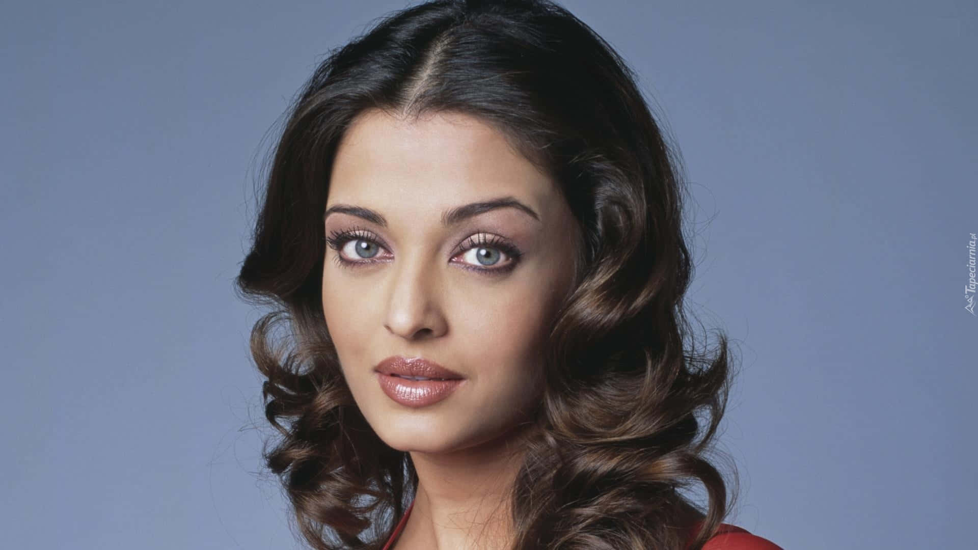 Aishwarya Rai 桌布 壁紙 — 一位深色波浪髮、眼神迷人的女性特寫肖像，背景為柔和的藍色。