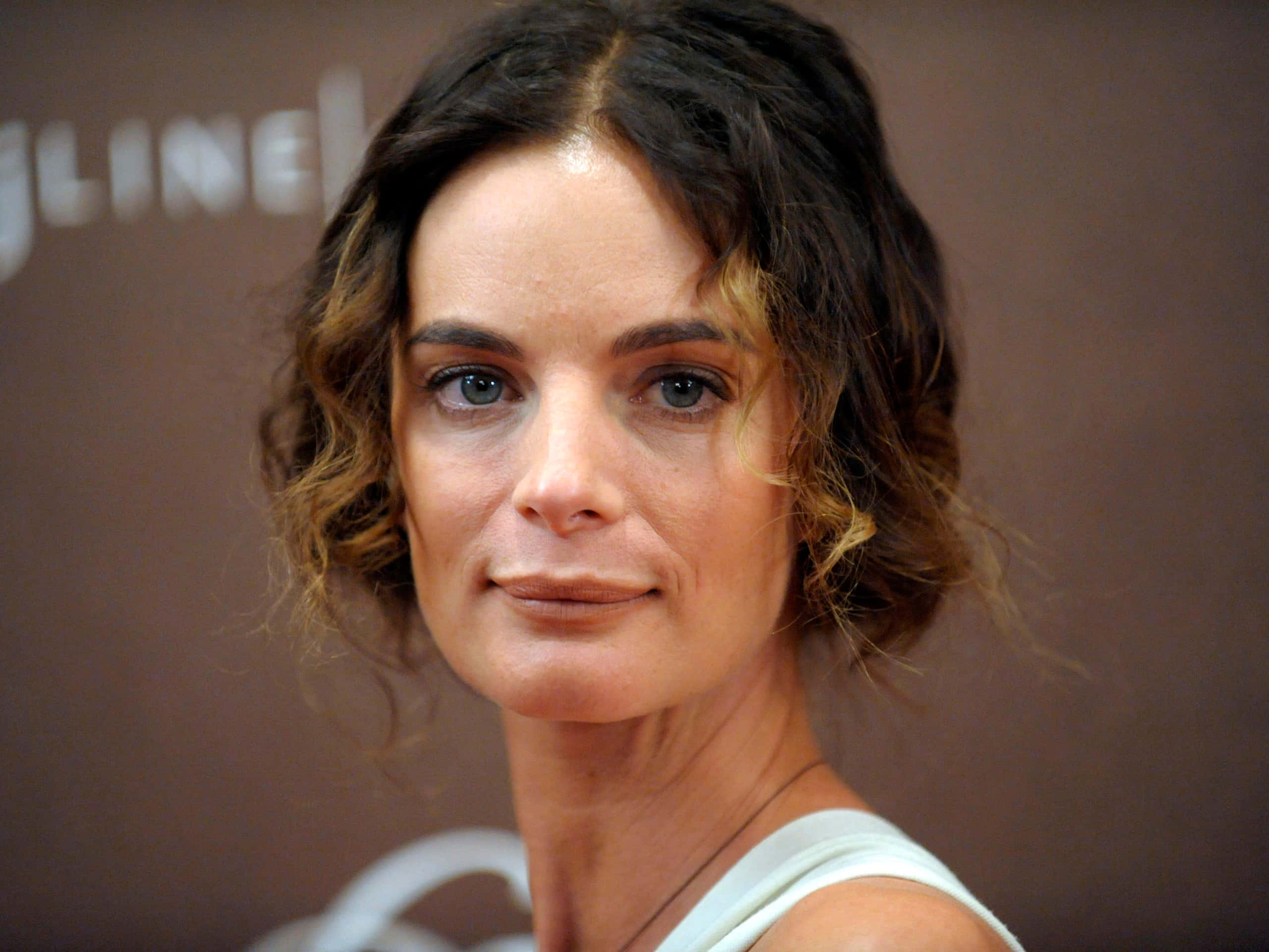 Gabrielle Anwar 桌布 壁紙 — 一位深色波浪髮、藍眼睛的女性穿著白色上衣，背景為模糊的棕色，特寫肖像。