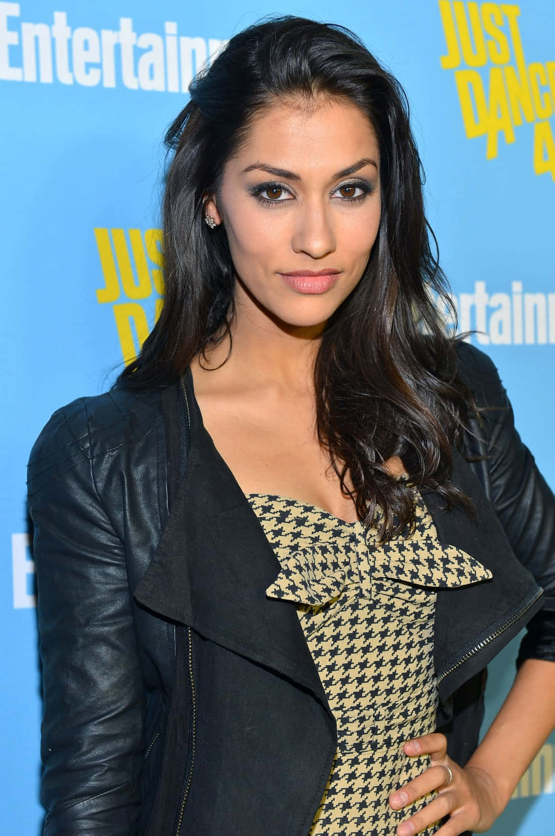 Janina Gavankar 桌布 壁紙 — 一位深色波浪髮的女性在「Just Dance」活動中穿著黑色皮褸配千鳥格連身裙。