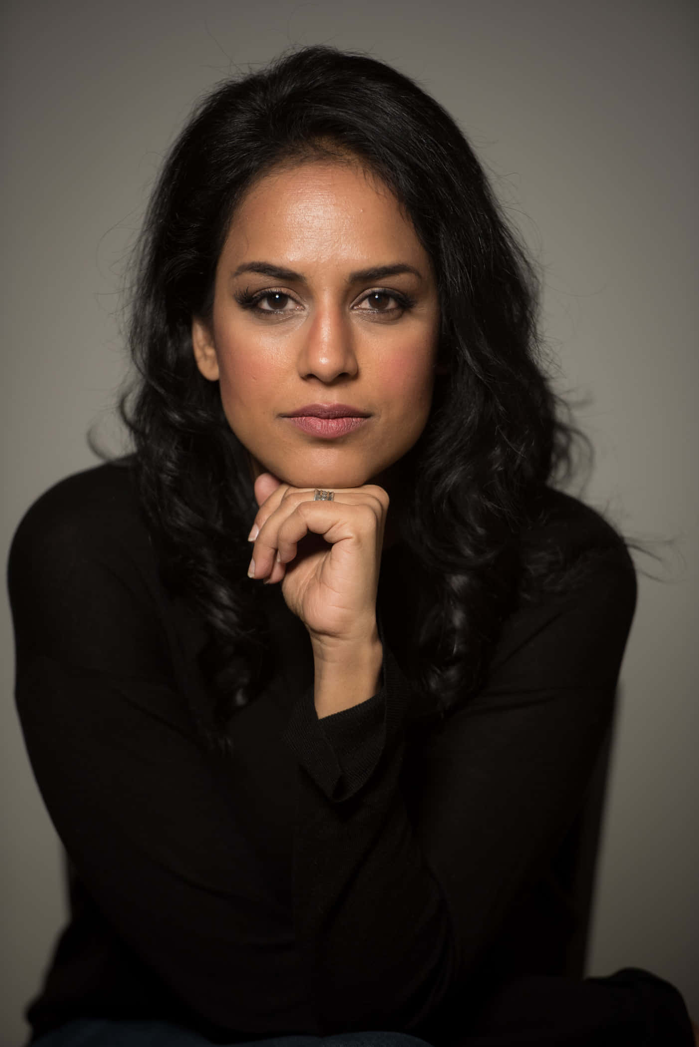 Agam Darshi 桌布 壁紙 — 一位深色波浪髮的女性在灰色中性背景上將下巴靠在手上。