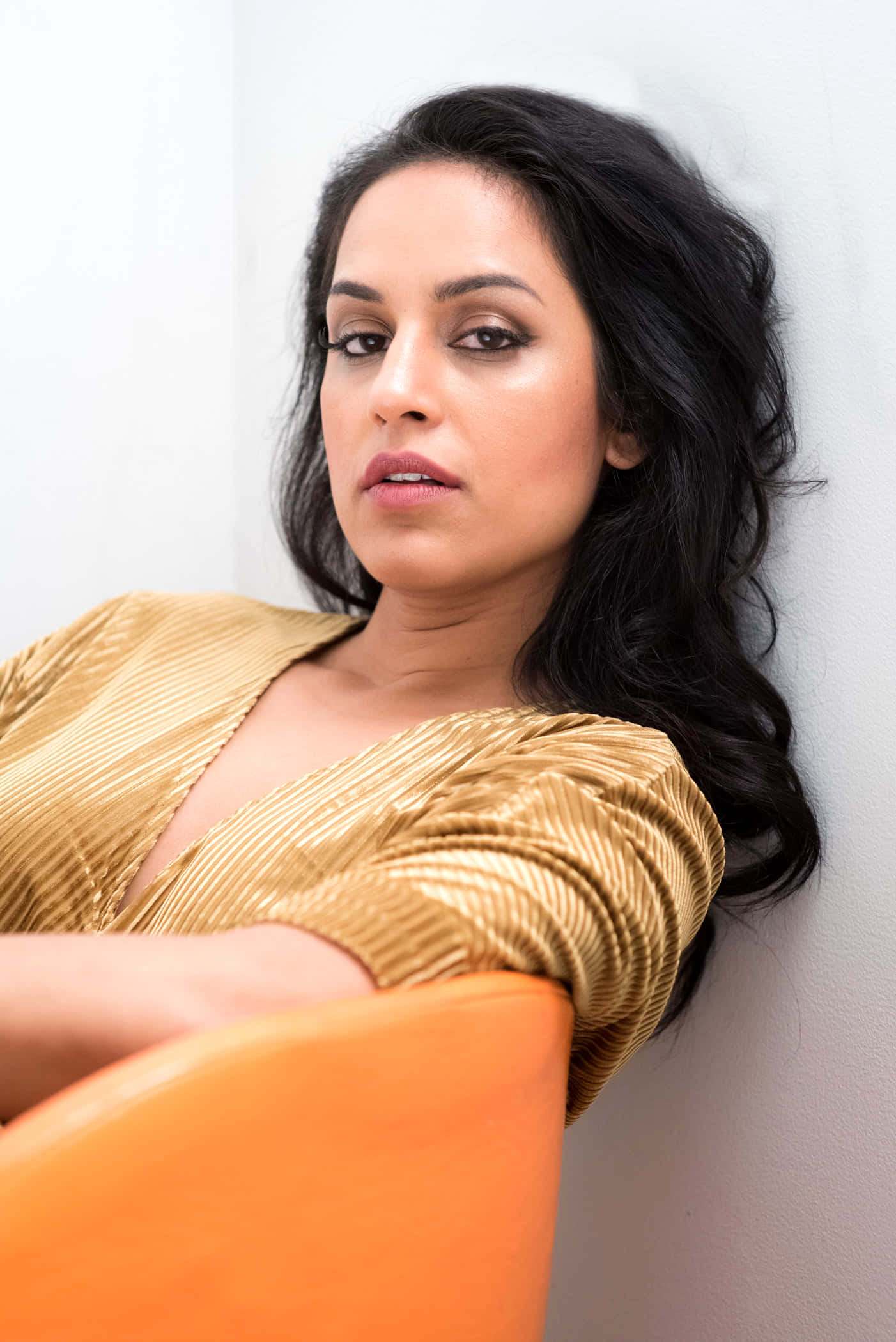 Agam Darshi 桌布 壁紙 — 一位深色波浪髮的女性穿著金色百褶上衣和橙色椅子靠在牆上。