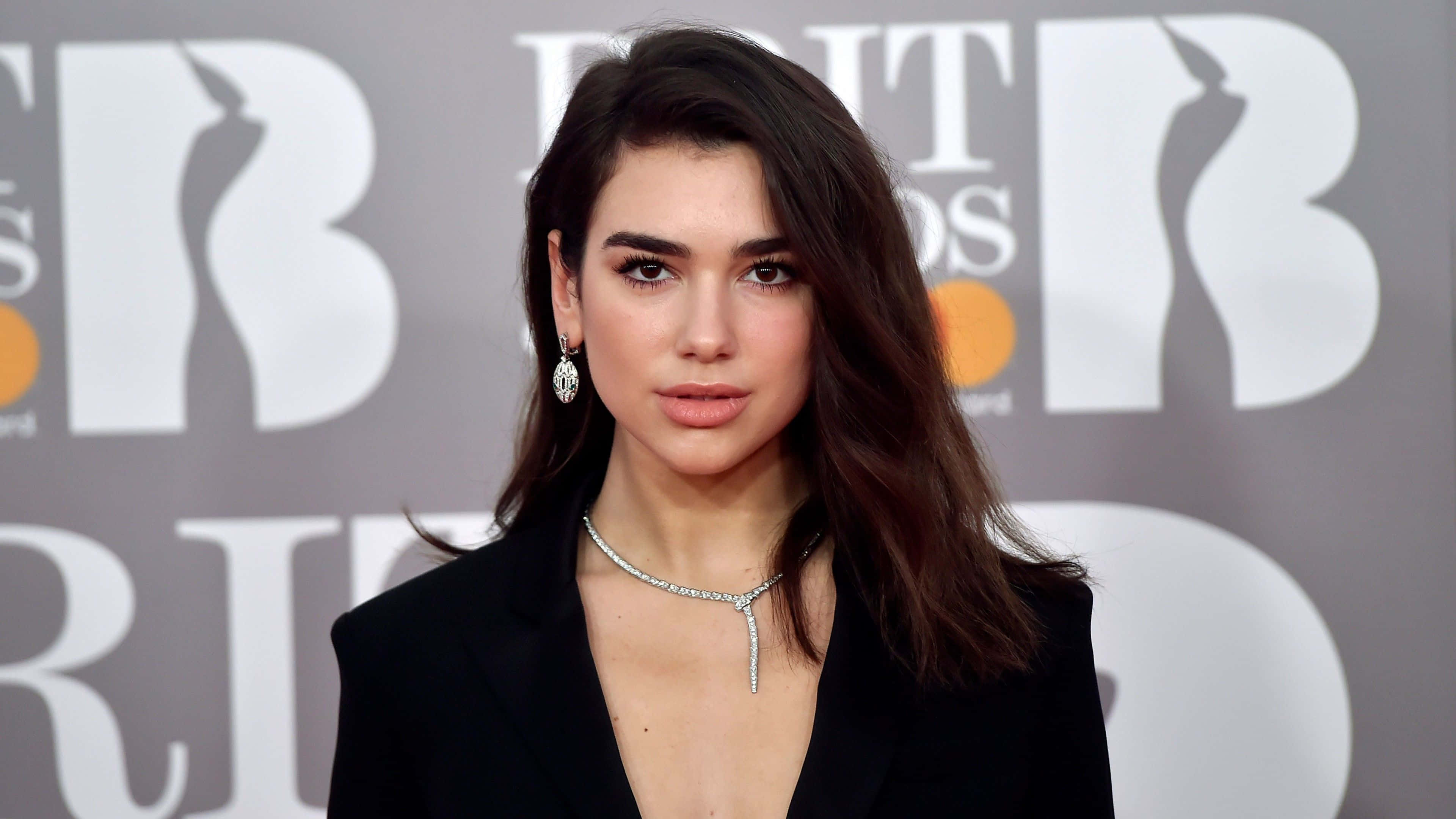 Dua Lipa 桌布 壁紙 — 一位深色頭髮、佩戴優雅珠寶的自信女性，在品牌背景前於紅地毯活動中自信地站立。