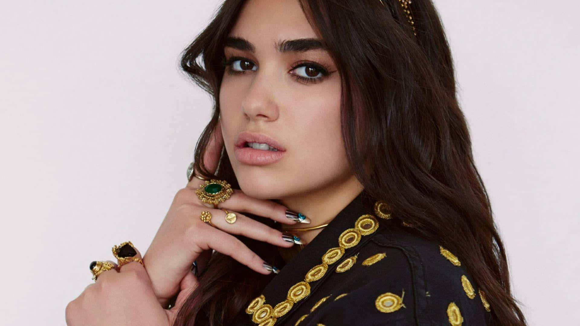 Dua Lipa 桌布 壁紙 — 一位深色頭髮、佩戴大膽珠寶的女性在純白背景前擺姿勢，手靠近臉部。