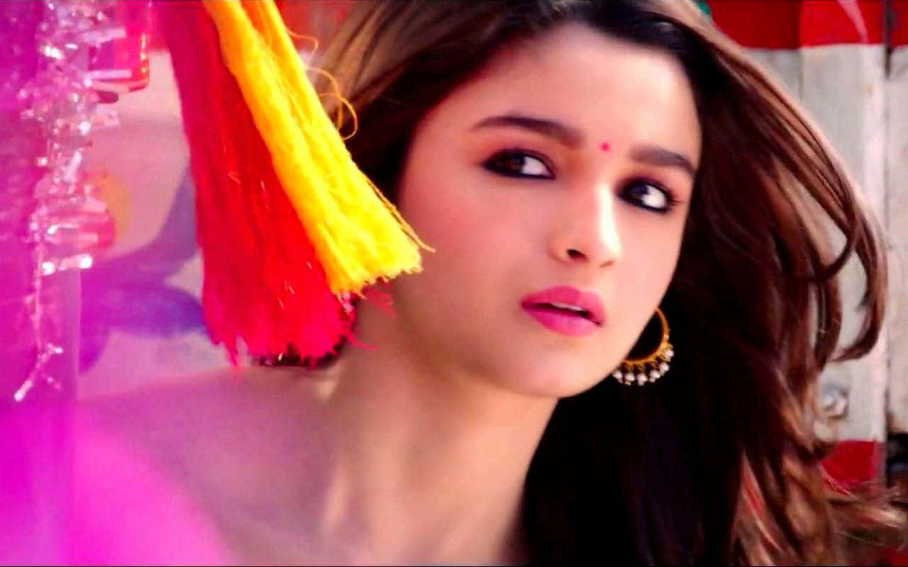 Alia Bhatt 桌布 壁紙 — 一位深色頭髮、塗著粉紅色唇膏的女性，在粉紅色燈光下，在鮮豔的紅色和黃色流蘇旁邊凝視著。