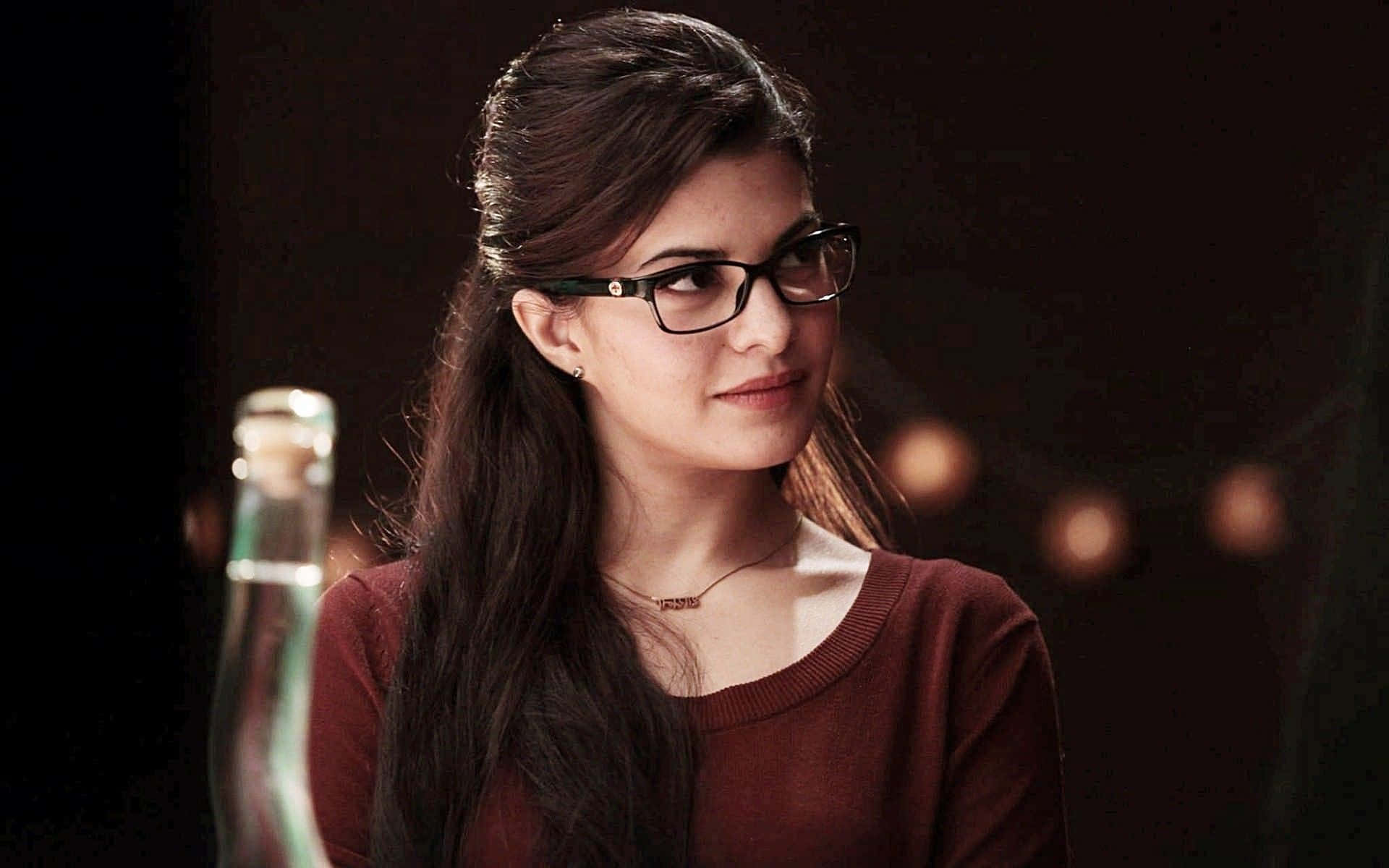 Jacqueline Fernandez 桌布 壁紙 — 一位深色頭髮、戴眼鏡的年輕女性，在昏暗的環境中穿著栗色毛衣，柔和地微笑，背景有溫暖的散景燈光。