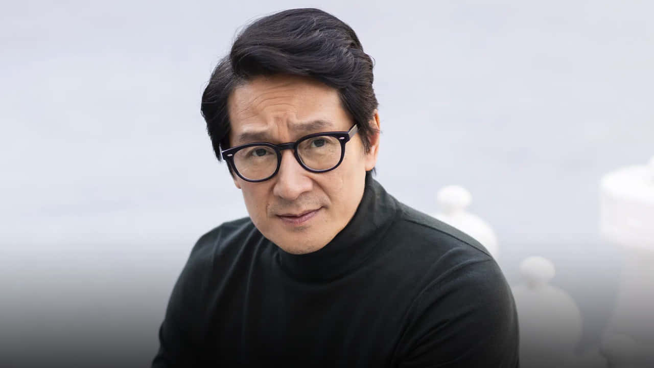 Ke Huy Quan 桌布 壁紙 — 一位深色頭髮、戴眼鏡的男子，身穿黑色高領毛衣，背景為柔和的灰色，帶有模糊的白色形狀。