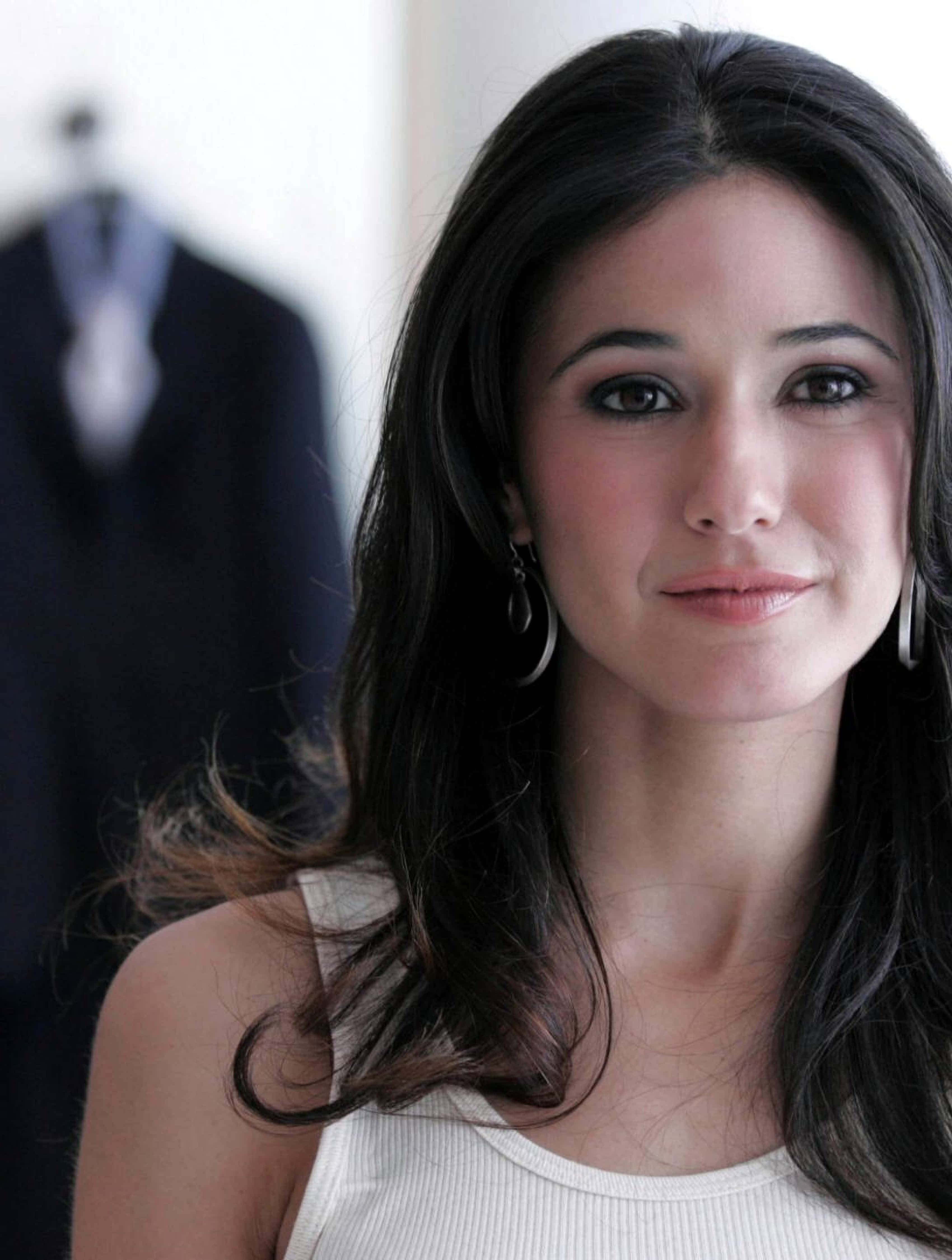 Emmanuelle Chriqui 桌布 壁紙 — 一位深色頭髮、戴著圈圈耳環的女性，穿著白色背心，背景中有一件模糊的西裝外套，她溫柔地微笑。