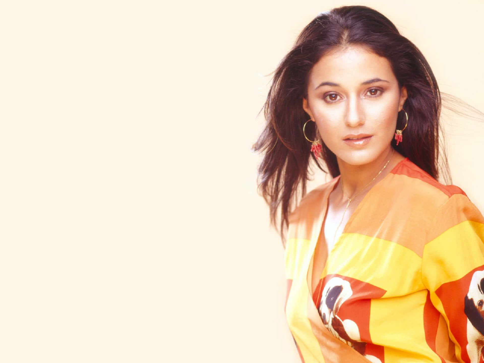 Emmanuelle Chriqui 桌布 壁紙 — 一位深色頭髮、戴著圈狀耳環的女性，在柔和的奶油色背景前穿著鮮豔的橙黃圖案上衣。