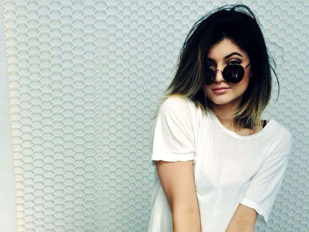 Kylie Jenner 桌布 壁紙 — 一位深色頭髮、戴著圓形太陽眼鏡的年輕女性，自信地站在白色六角形瓷磚牆前擺姿勢。
