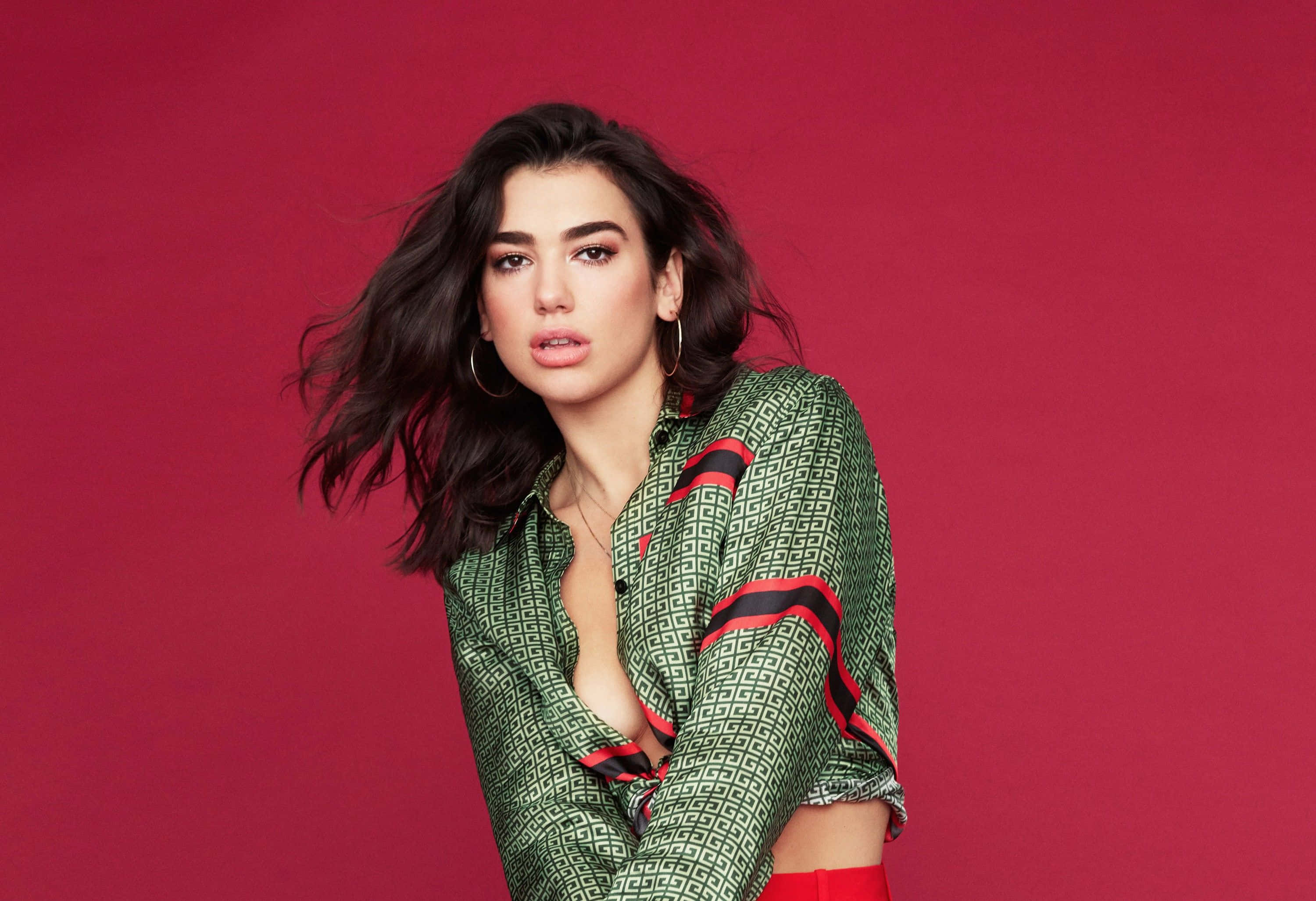 Dua Lipa 桌布 壁紙 — 一位深色頭髮、戴著圓環耳環的女性，穿著綠色圖案襯衫，在鮮紅色背景前自信地擺姿勢。