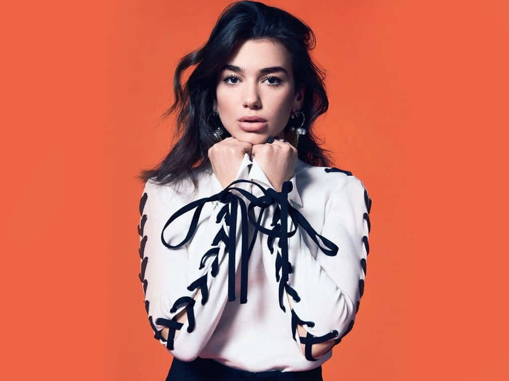 Dua Lipa 桌布 壁紙 — 一位深色頭髮、戴著大膽耳環的女性，在充滿活力的橙色背景前穿著白色繫帶上衣擺姿勢。