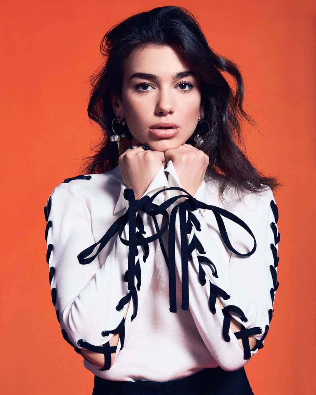 Dua Lipa 桌布 壁紙 — 一位深色頭髮、戴著大膽耳環的女性，穿著帶有黑色繫帶細節的白色襯衫，在充滿活力的橙色背景下直視鏡頭。