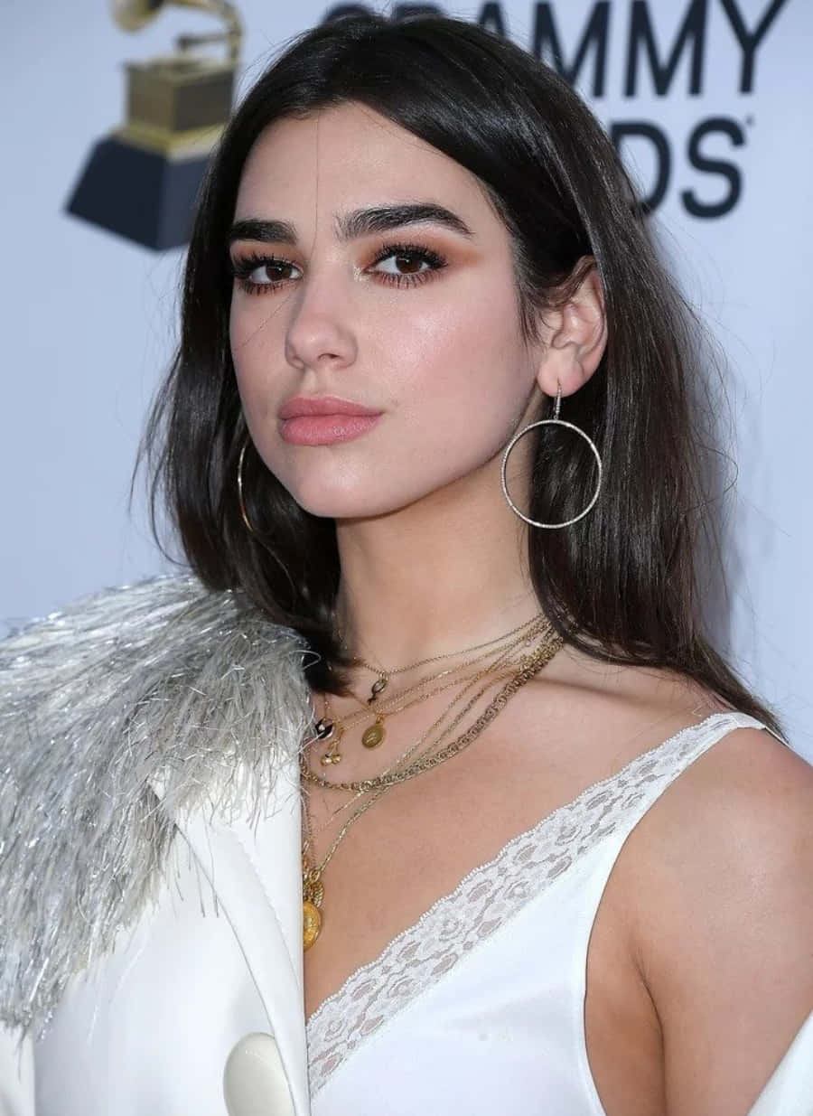 杜娃·利帕 桌布 壁紙 — 一位深色頭髮、戴著金色圓環耳環的迷人女性在格林美獎活動中穿著白色蕾絲上衣和羽毛外套擺姿勢。