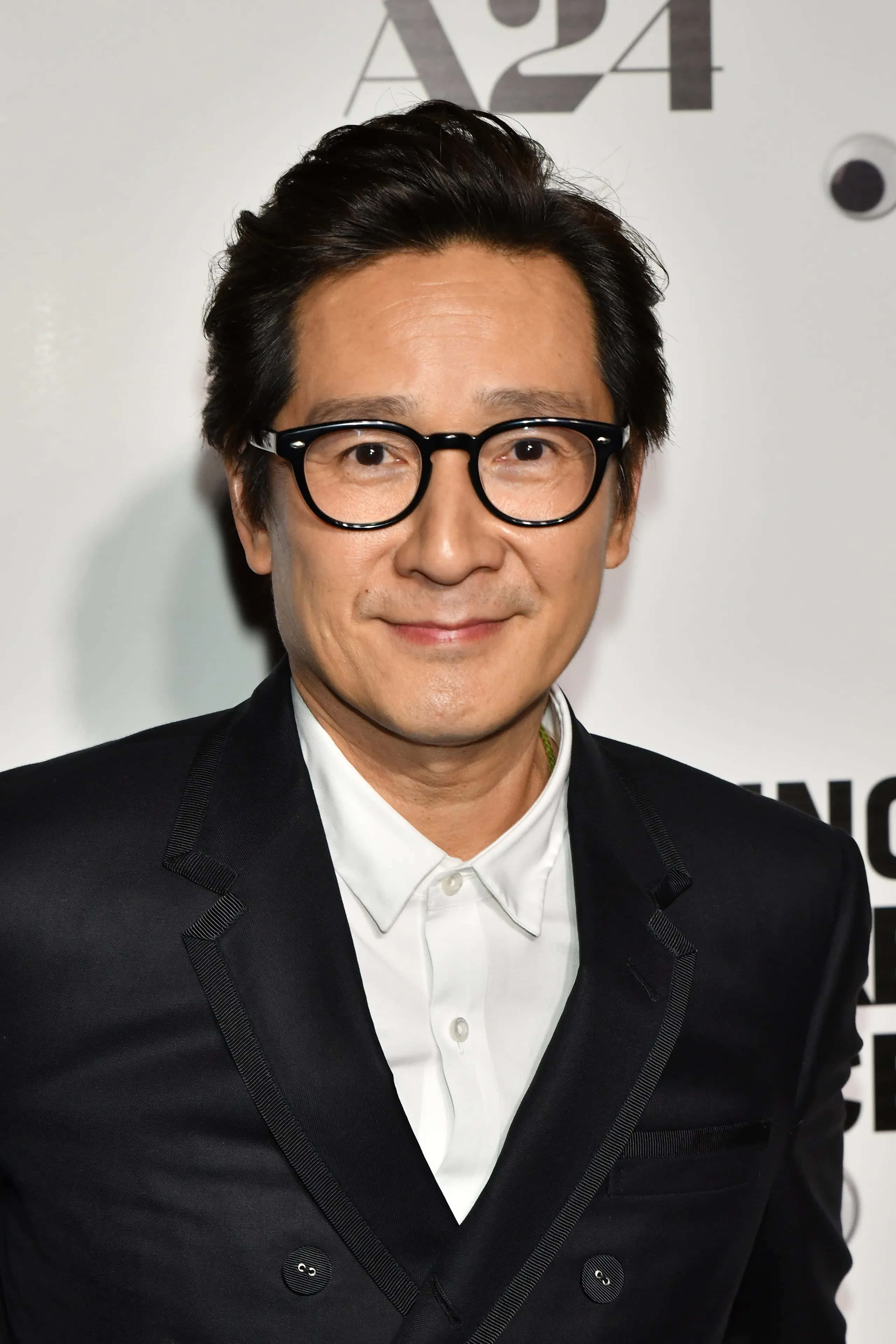 Ke Huy Quan 桌布 壁紙 — 一位深色頭髮、戴黑色框眼鏡的男士，身穿黑色西裝和白色襯衫，在品牌背景前溫暖地微笑。