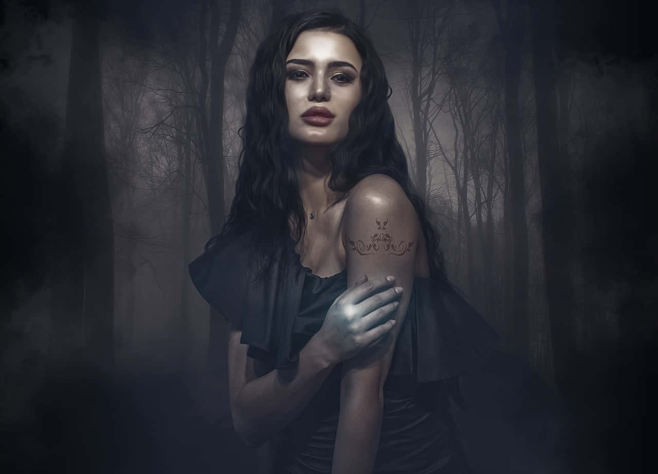 Gothic Fashion 桌布 壁紙 — 一位深色頭髮、手臂有紋身的女性，站在霧氣瀰漫、陰暗的森林中，周圍是光禿禿的樹木。