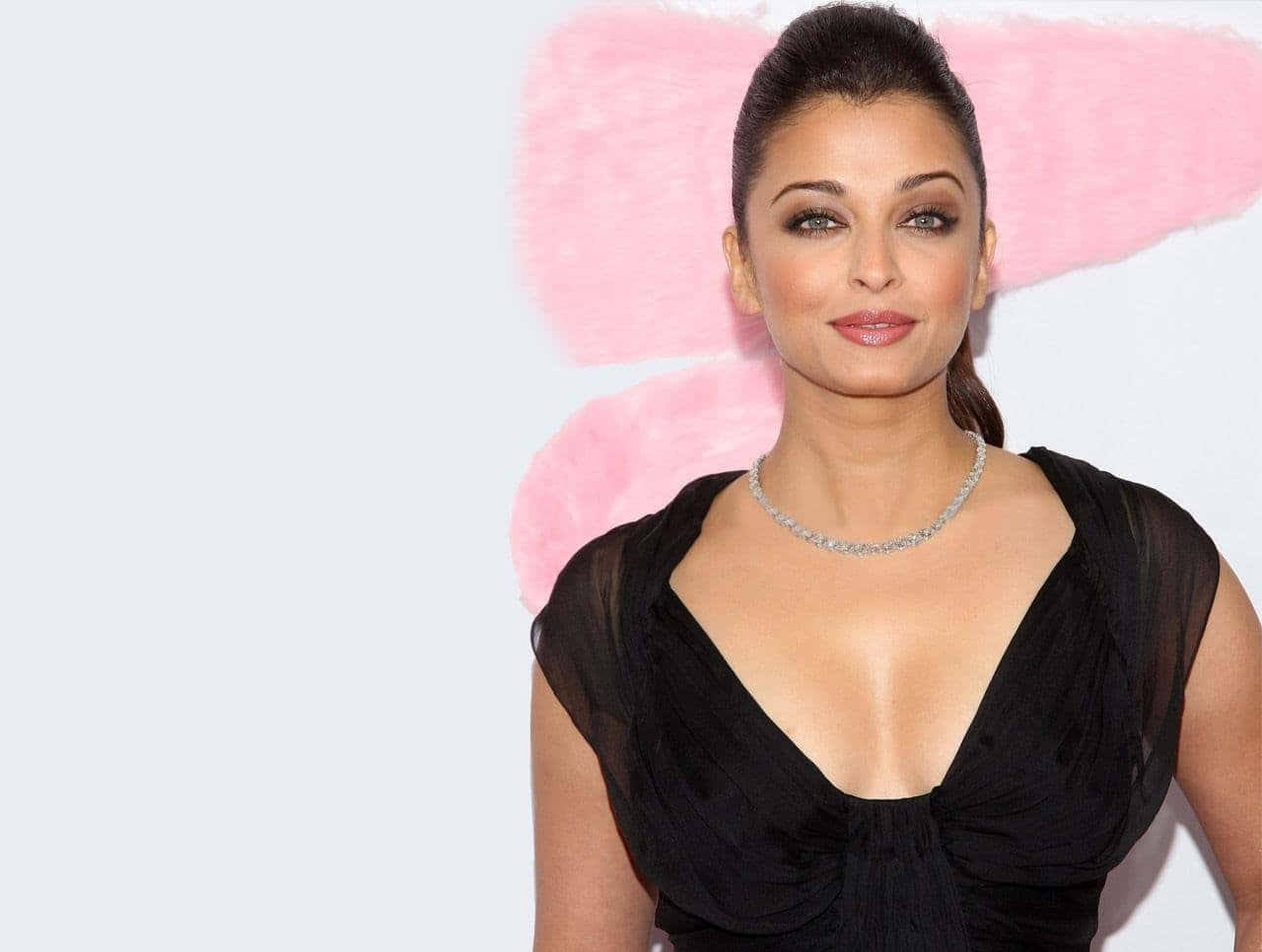 Aishwarya Rai 桌布 壁紙 — 一位深色頭髮、梳著光滑高髻的女性，穿著黑色連衣裙和鑽石項鍊，背景是柔和的粉紅色和白色。