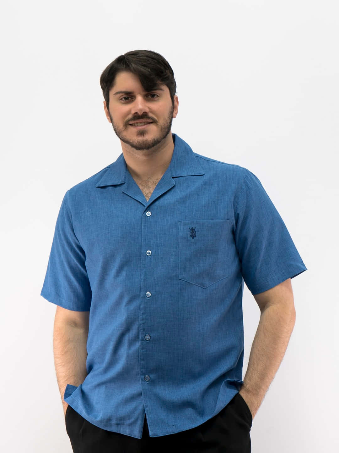 Denim Blue 桌布 壁紙 — 一位深色頭髮、留鬍鬚的男子，身穿藍色短袖鈕扣襯衫（有口袋），在純白背景前對著鏡頭微笑。