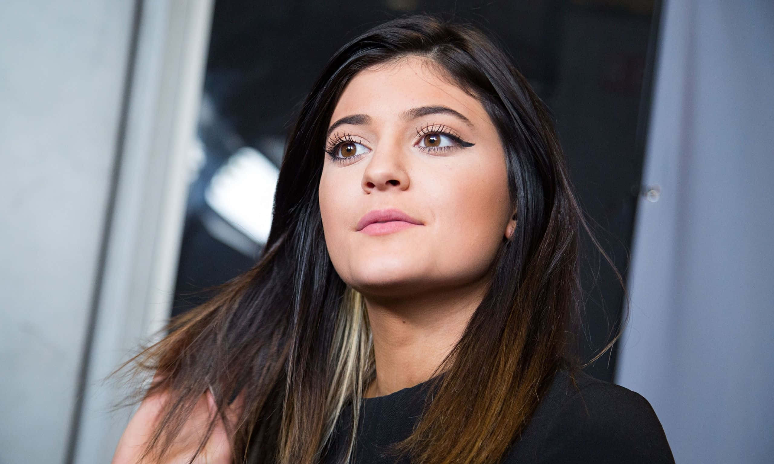 Kylie Jenner 桌布 壁紙 — 一位深色頭髮、畫著粗眼線的女性，在模糊的現代背景前若有所思地向上凝視的特寫肖像。