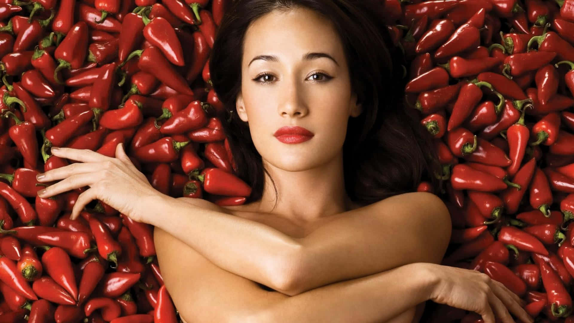 maggie q 桌布 壁紙 — 一位深色頭髮、紅唇的女性躺在鮮紅色的辣椒中，雙臂交叉在胸前。