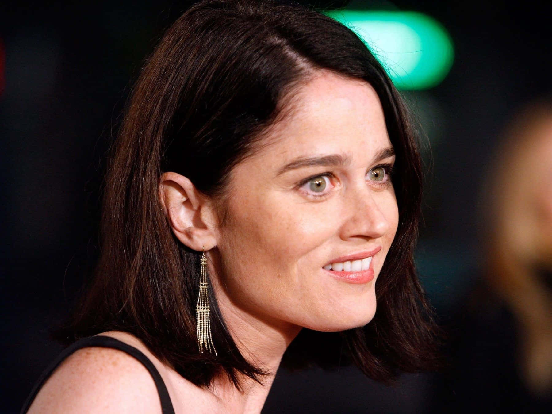 Robin Tunney 桌布 壁紙 — 一位深色頭髮、綠色眼睛的女性，在模糊的背景前戴著搖晃的金色耳環，溫柔地微笑。