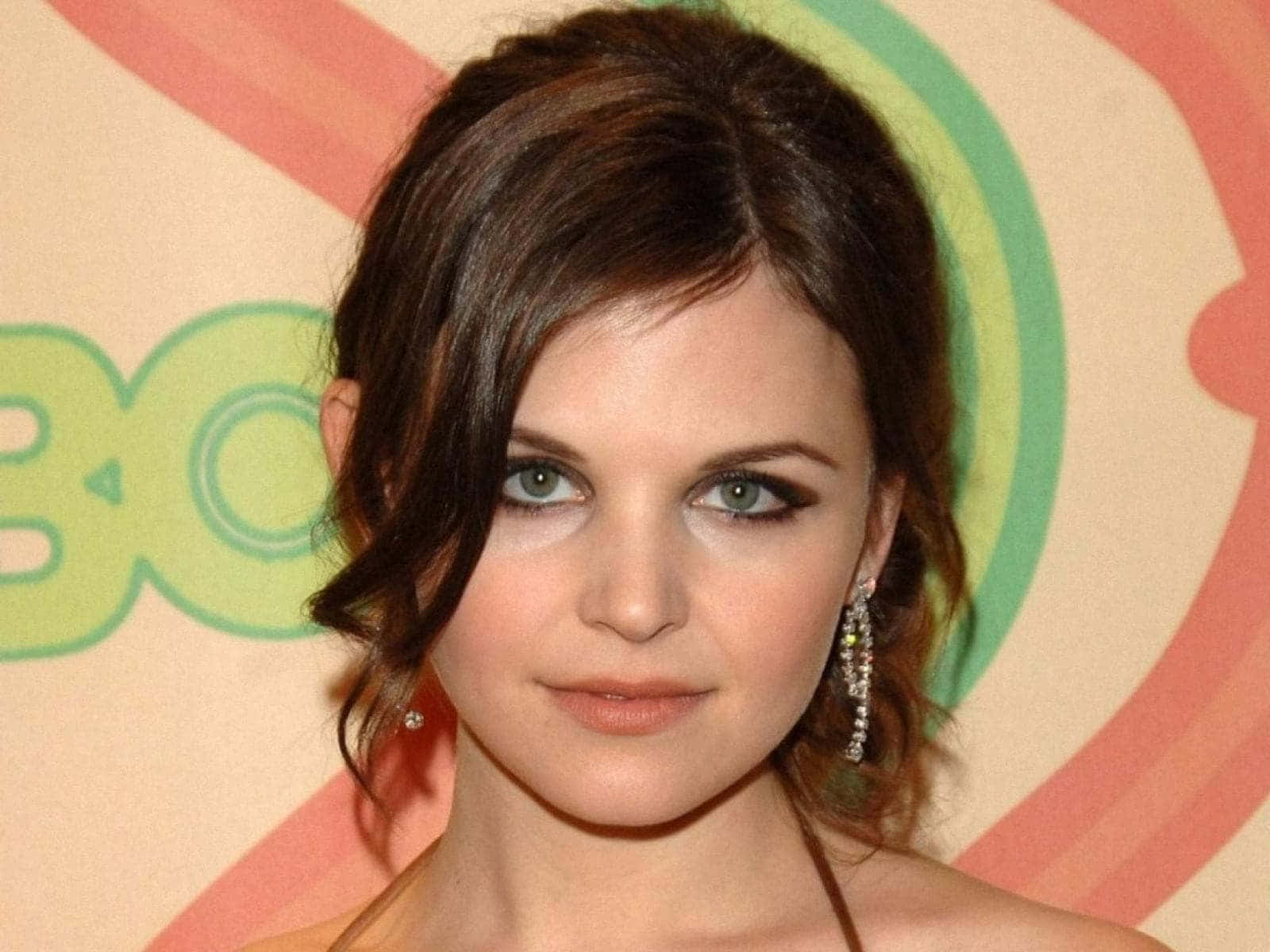 ginnifer goodwin 桌布 壁紙 — 一位深色頭髮、綠色眼睛的女性戴著閃閃發光的耳環，以色彩繽紛的抽象背景為襯托的特寫肖像。