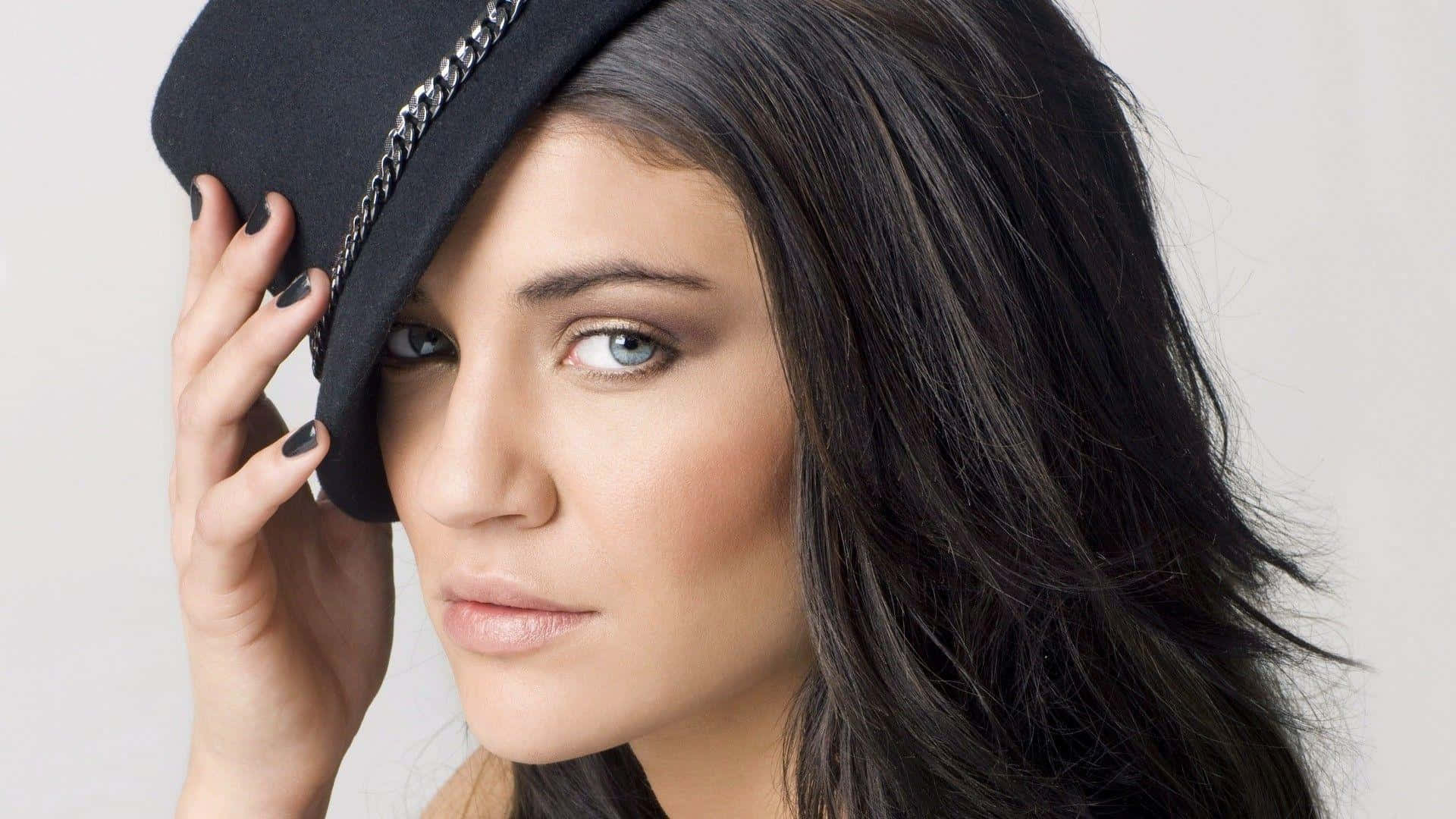 jessica szohr 桌布 壁紙 — 一位深色頭髮、藍色眼睛的女性，在純白色背景前拿著一頂帶有鏈條飾邊的黑色帽子。