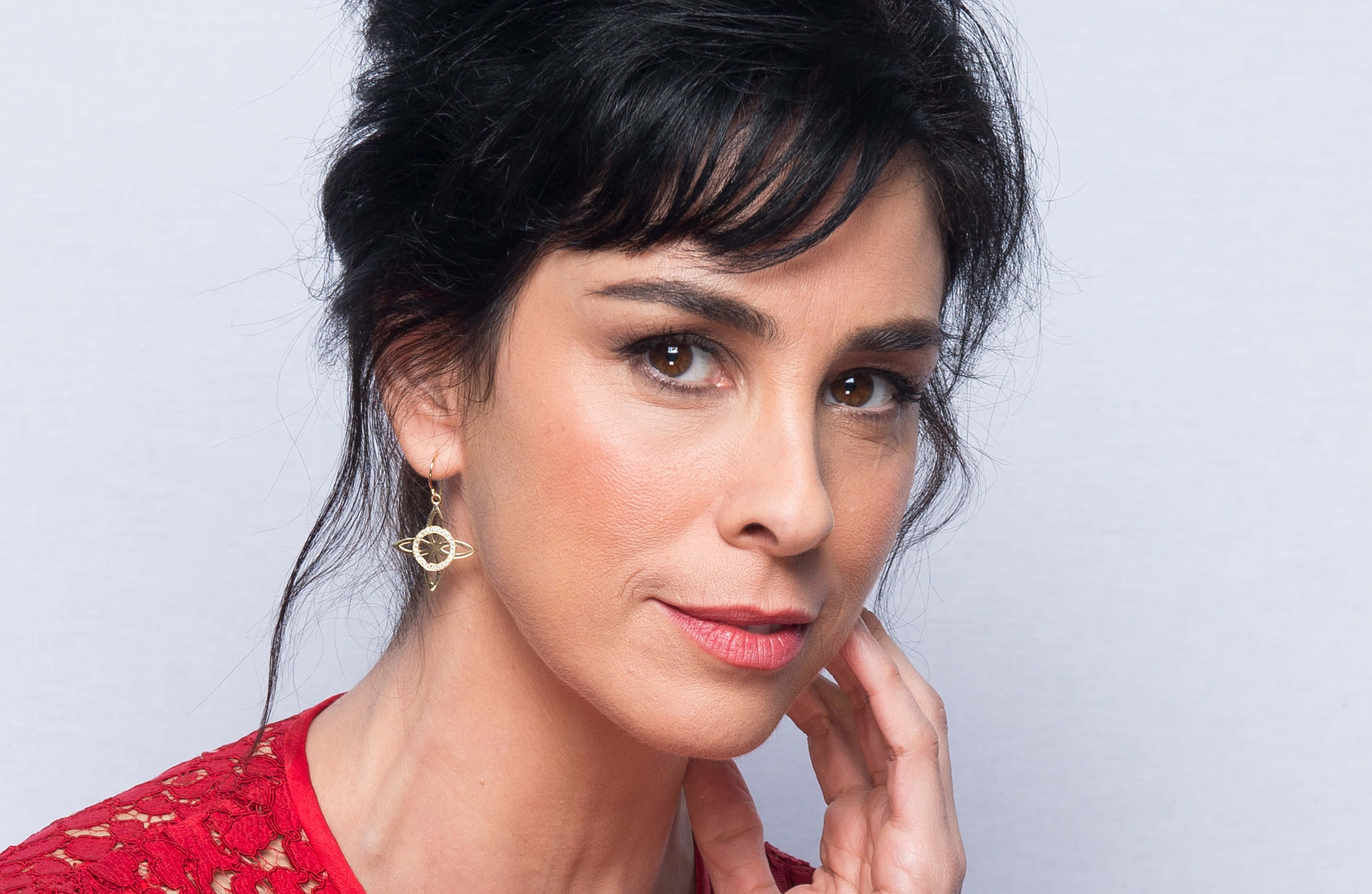 sarah silverman 桌布 壁紙 — 一位深色頭髮、身穿紅色蕾絲上衣的女性，在純灰色背景前輕觸臉頰，溫柔地凝視。