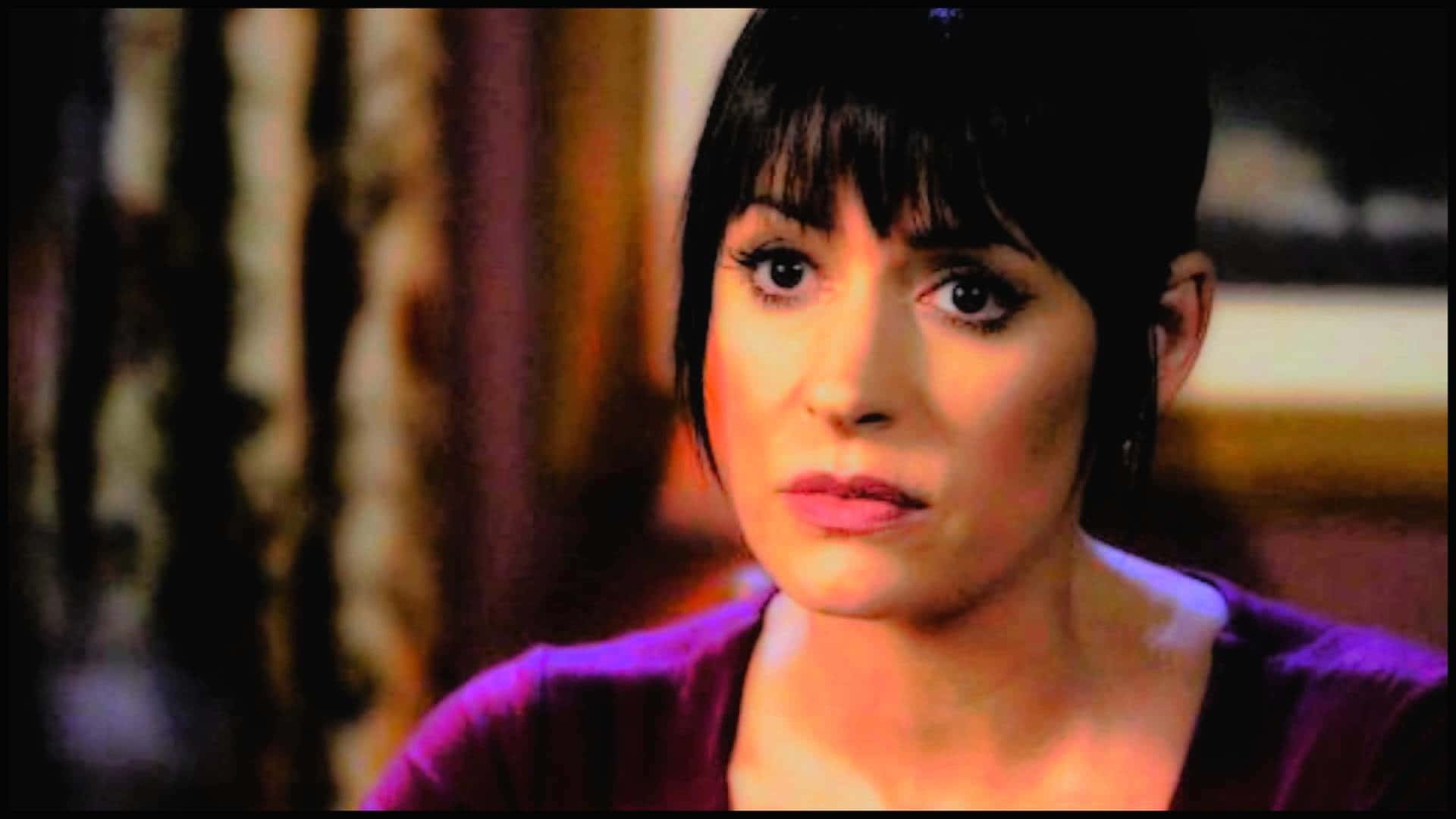 paget brewster 桌布 壁紙 — 一位深色頭髮、身穿紫色上衣的女性在溫暖的室內環境中凝視著。