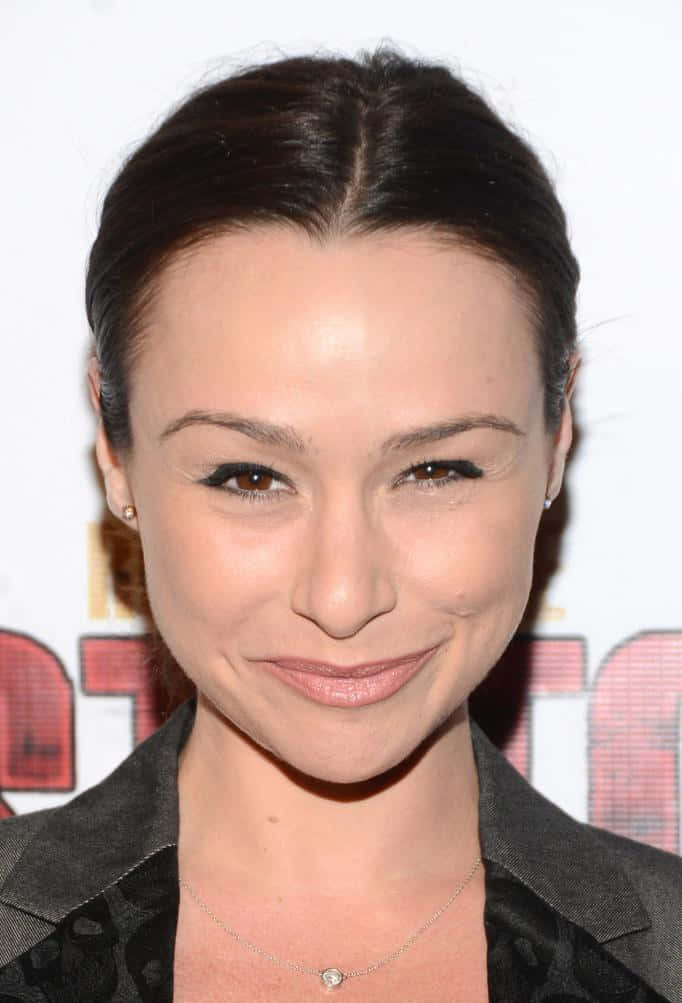 Danielle Harris 桌布 壁紙 — 一位深色頭髮向後梳的女性特寫肖像，她穿著深色西裝外套和精緻項鍊，對著鏡頭溫柔微笑。