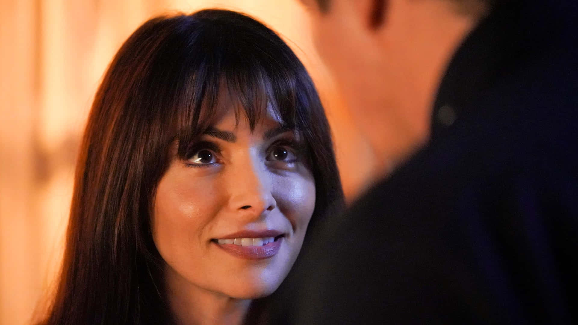sarah shahi 桌布 壁紙 — 一位深色頭髮和瀏海的女性在溫暖照明的室內環境中，微笑著看著鏡頭外的某人。